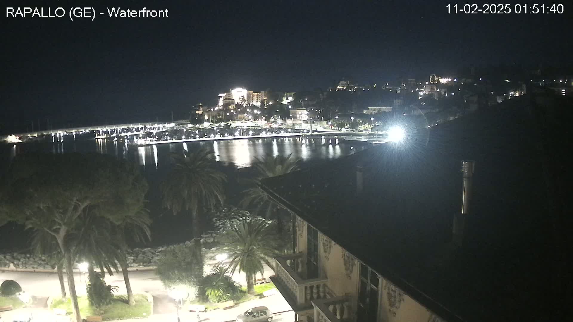A nighttime view of a harbor town's waterfront, showing a calm body of water reflecting city lights under a clear, dark sky.