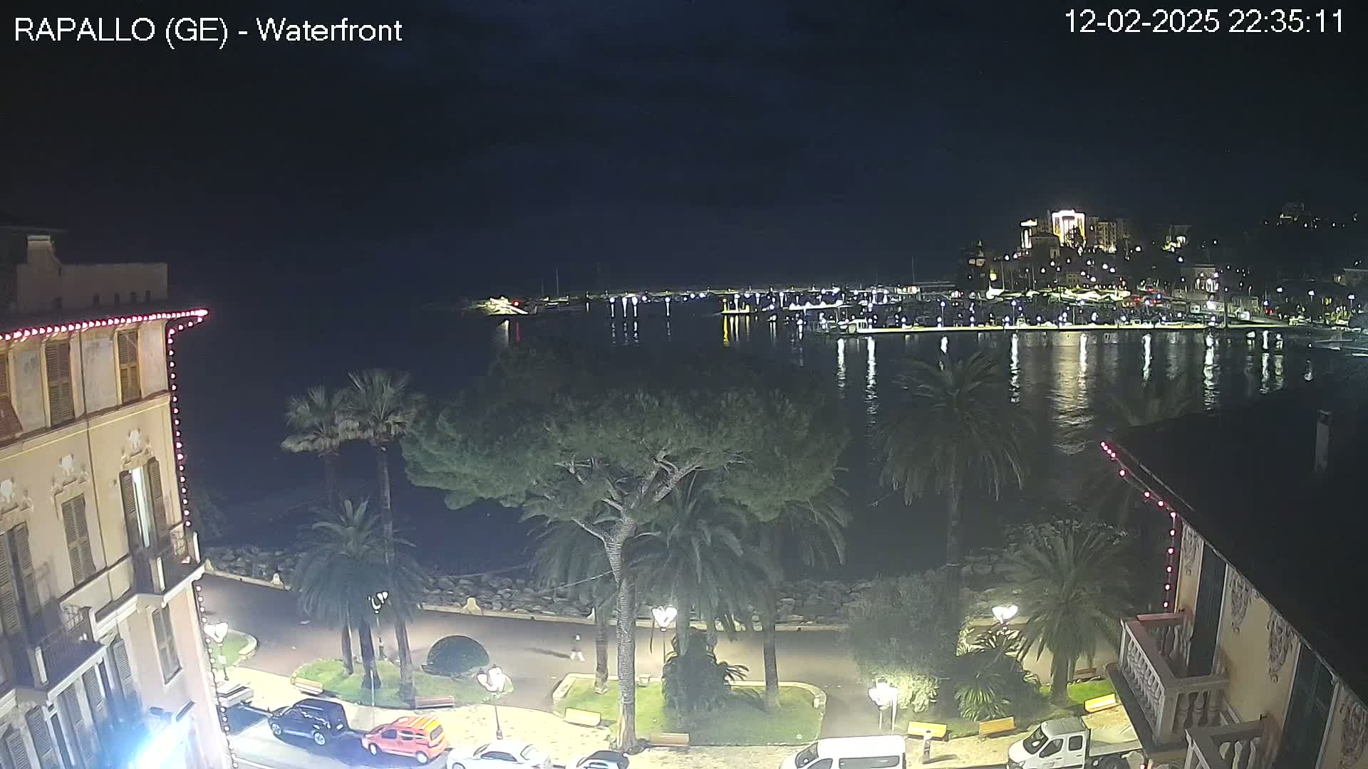 A nighttime view of a calm harbor, lined with boats and buildings, seen from a waterfront vantage point with palm trees and a building adorned with lights.
