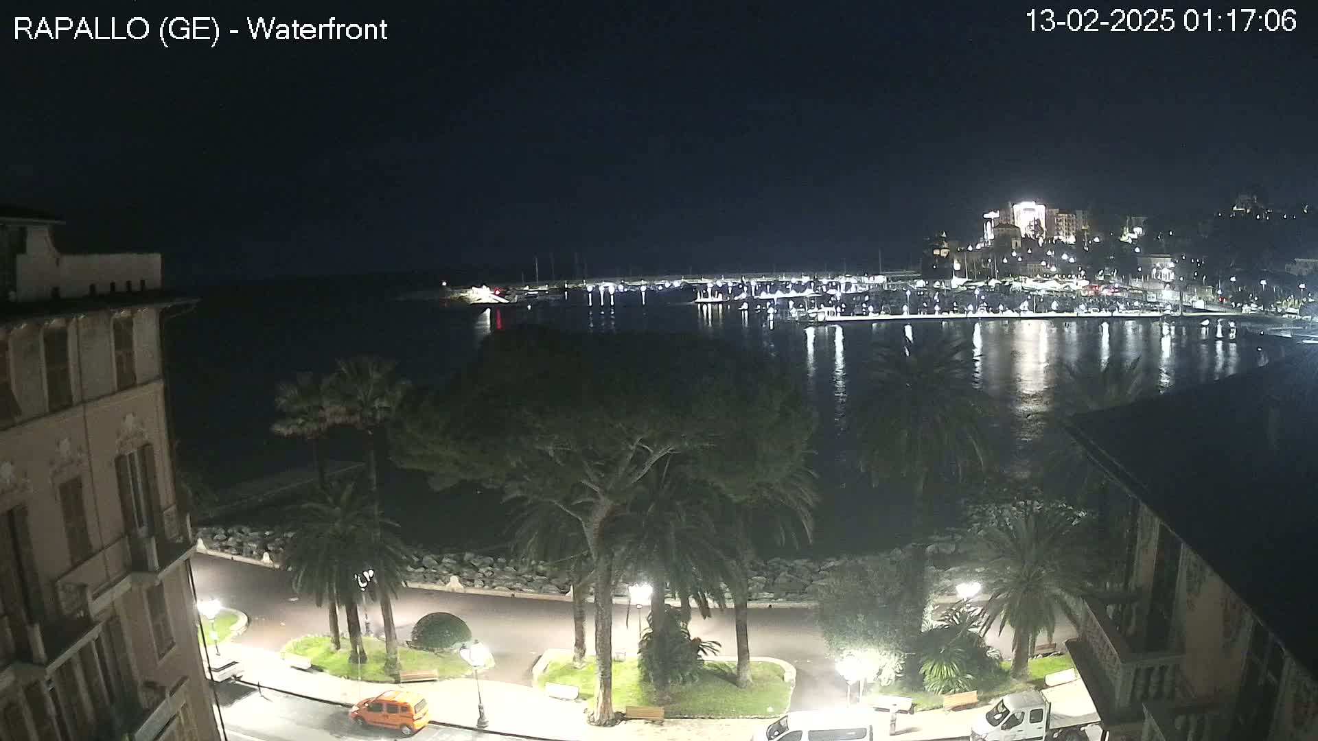 A nighttime view of a harbor filled with boats, lit buildings across the water, and a palm tree-lined waterfront promenade with a few cars parked along the side.