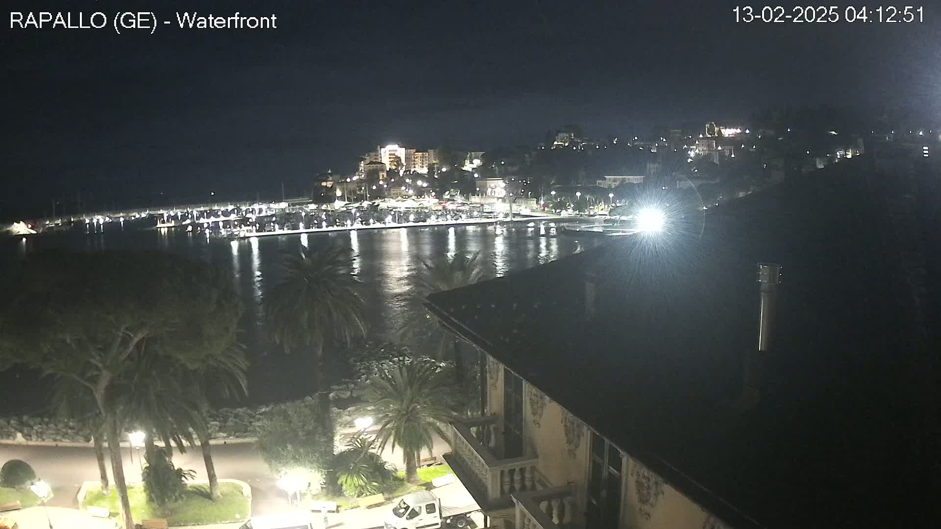 A nighttime view of a harbor, with boats moored and city lights illuminating the waterfront and hills beyond, under a clear sky.