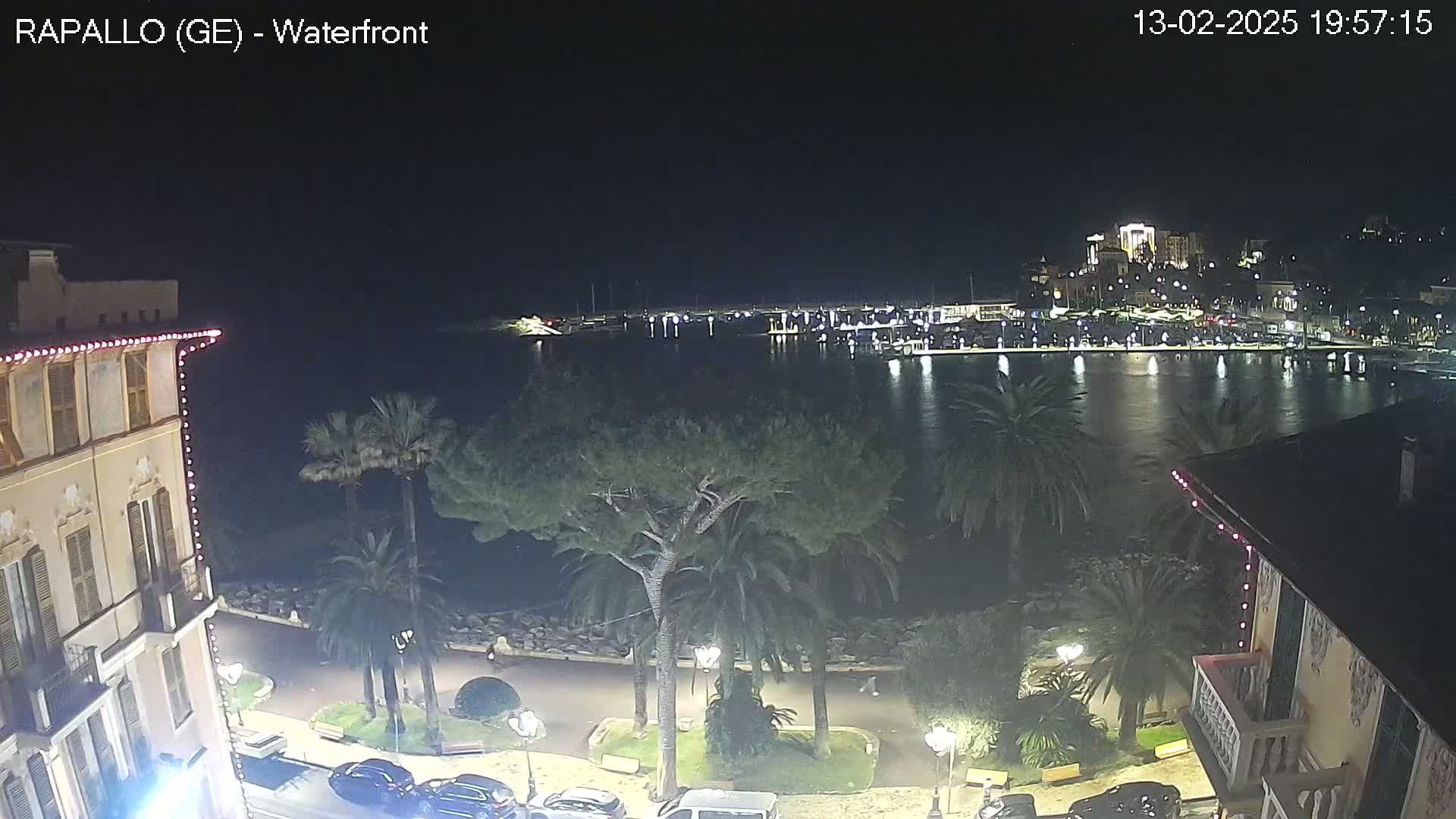 A nighttime view of a waterfront town, showing a calm harbor with boats, palm trees lining a paved walkway, and buildings with lights.