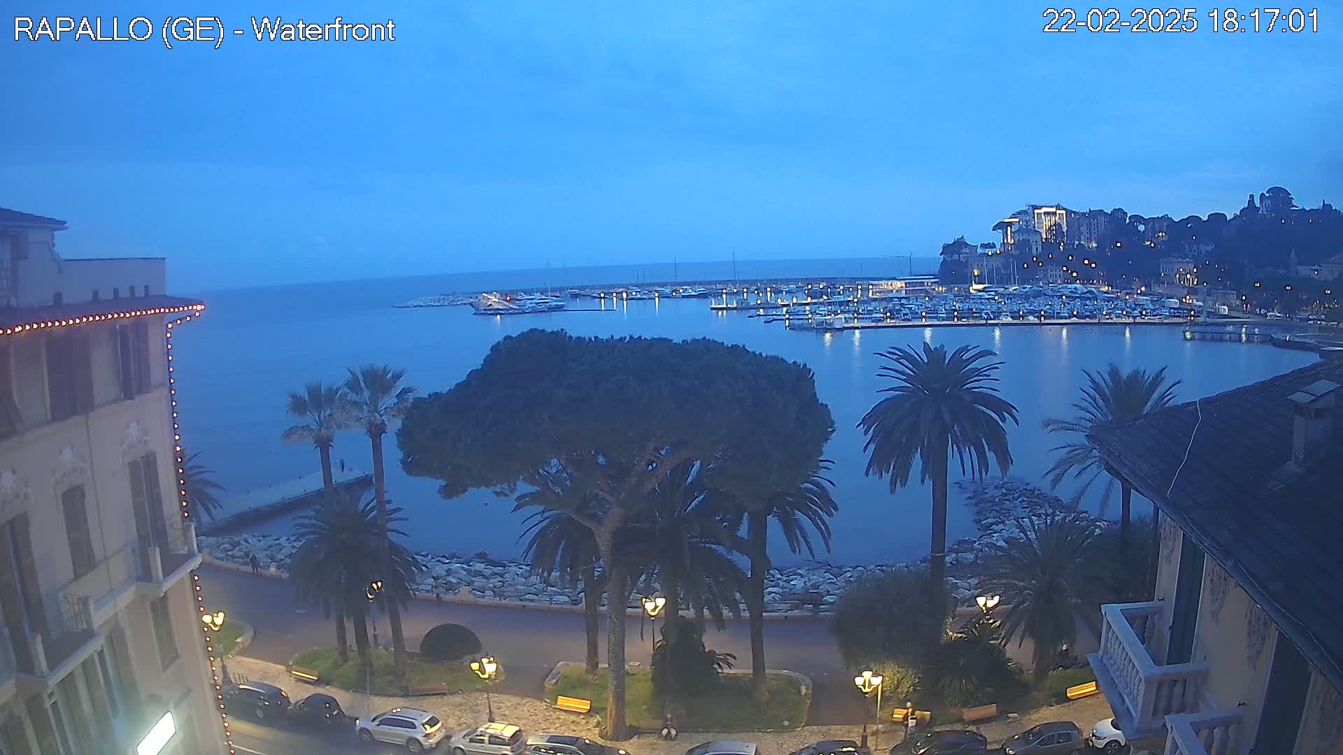 A waterfront town at dusk, showing a harbor filled with boats, palm trees lining a stone walkway, and buildings with lights illuminating the scene under a clear, twilight sky.