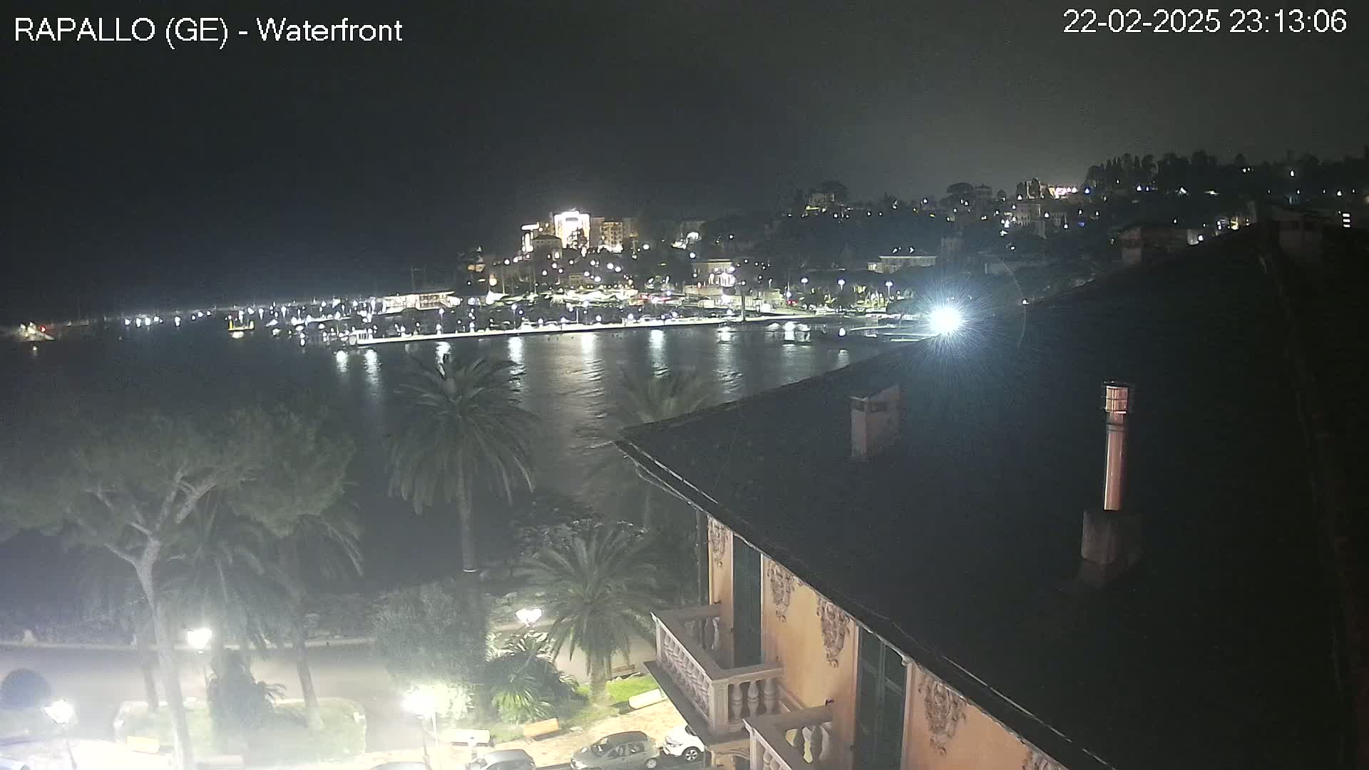 A nighttime view of a calm harbor with a town illuminated on a hill overlooking it, seen from a building's rooftop.