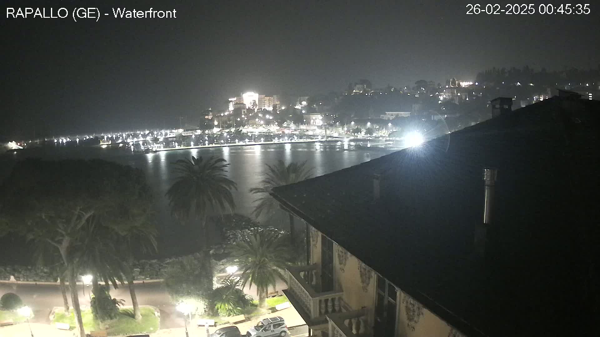 A nighttime view of a waterfront town, illuminated with lights, seen from a building's rooftop overlooking a calm body of water.