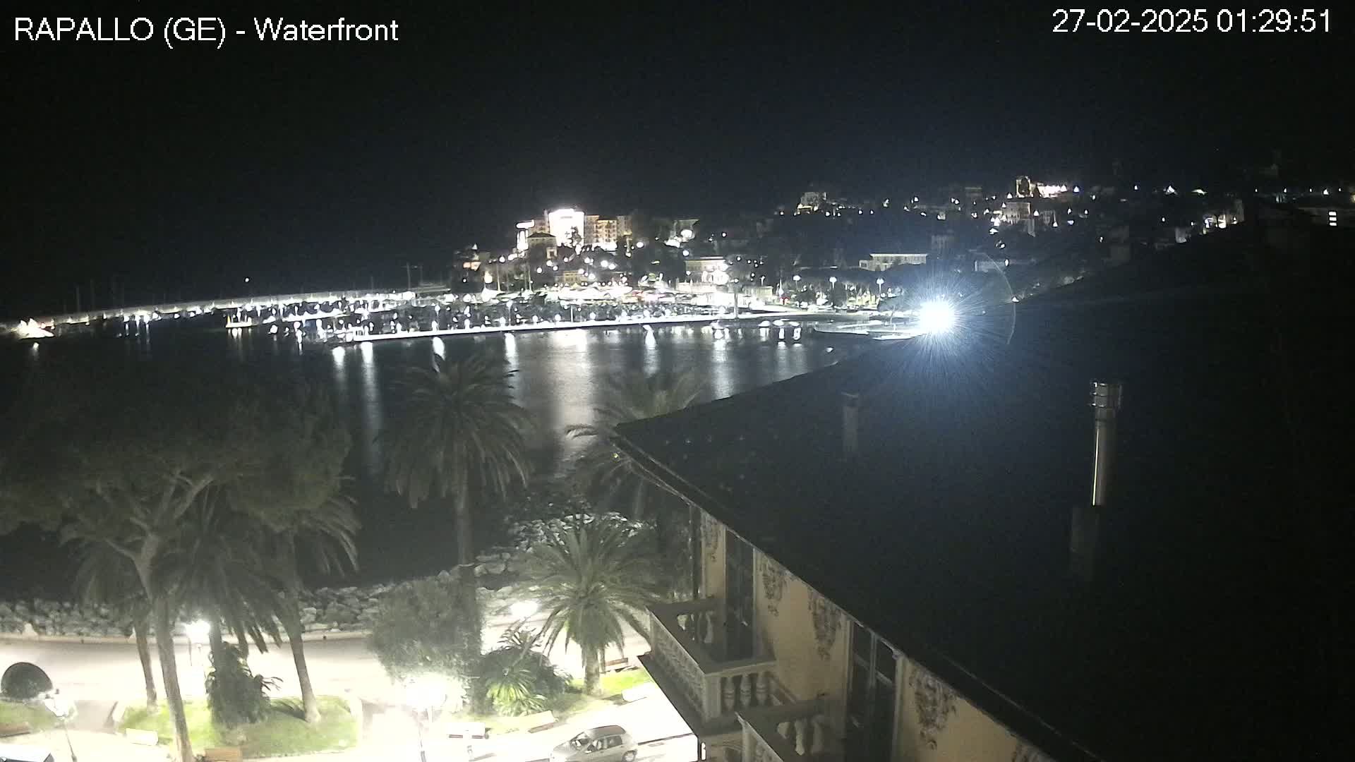 A nighttime view of a calm harbor with a town's lights reflecting on the water, palm trees lining a waterfront promenade, and a building's roof in the foreground.