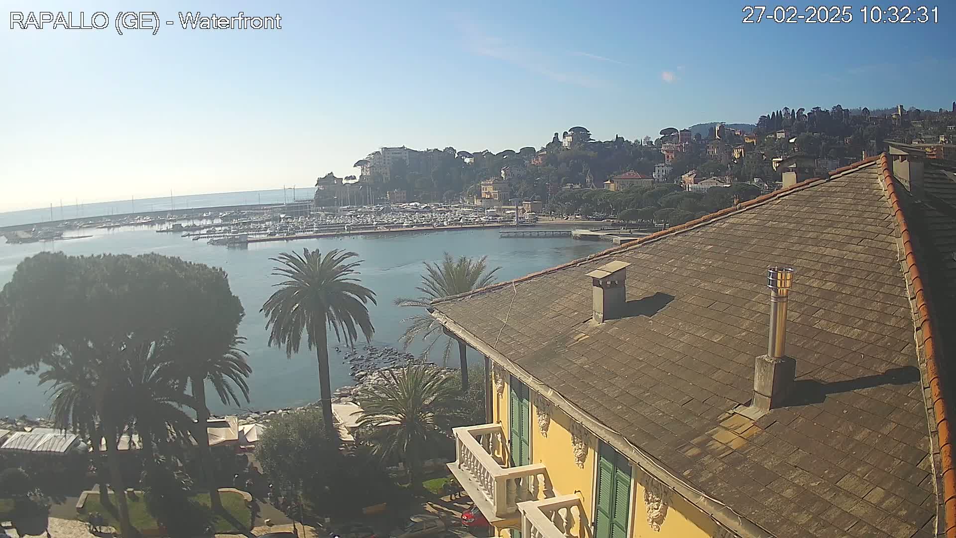 A clear, sunny day overlooks a waterfront harbor filled with boats and a hillside town, viewed from the tiled roof of a yellow building with green shutters.