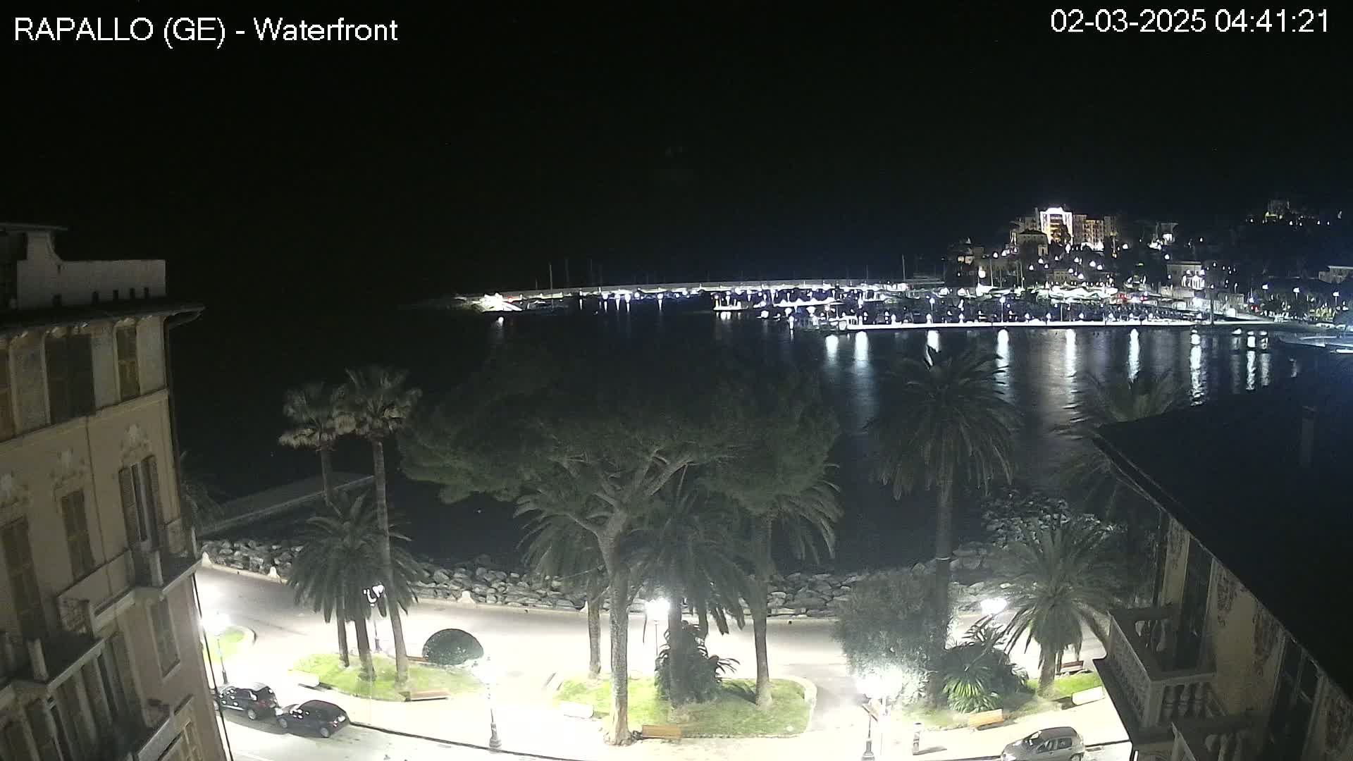 A nighttime view of a calm harbor, lined with buildings and palm trees, from a vantage point overlooking a waterfront promenade.
