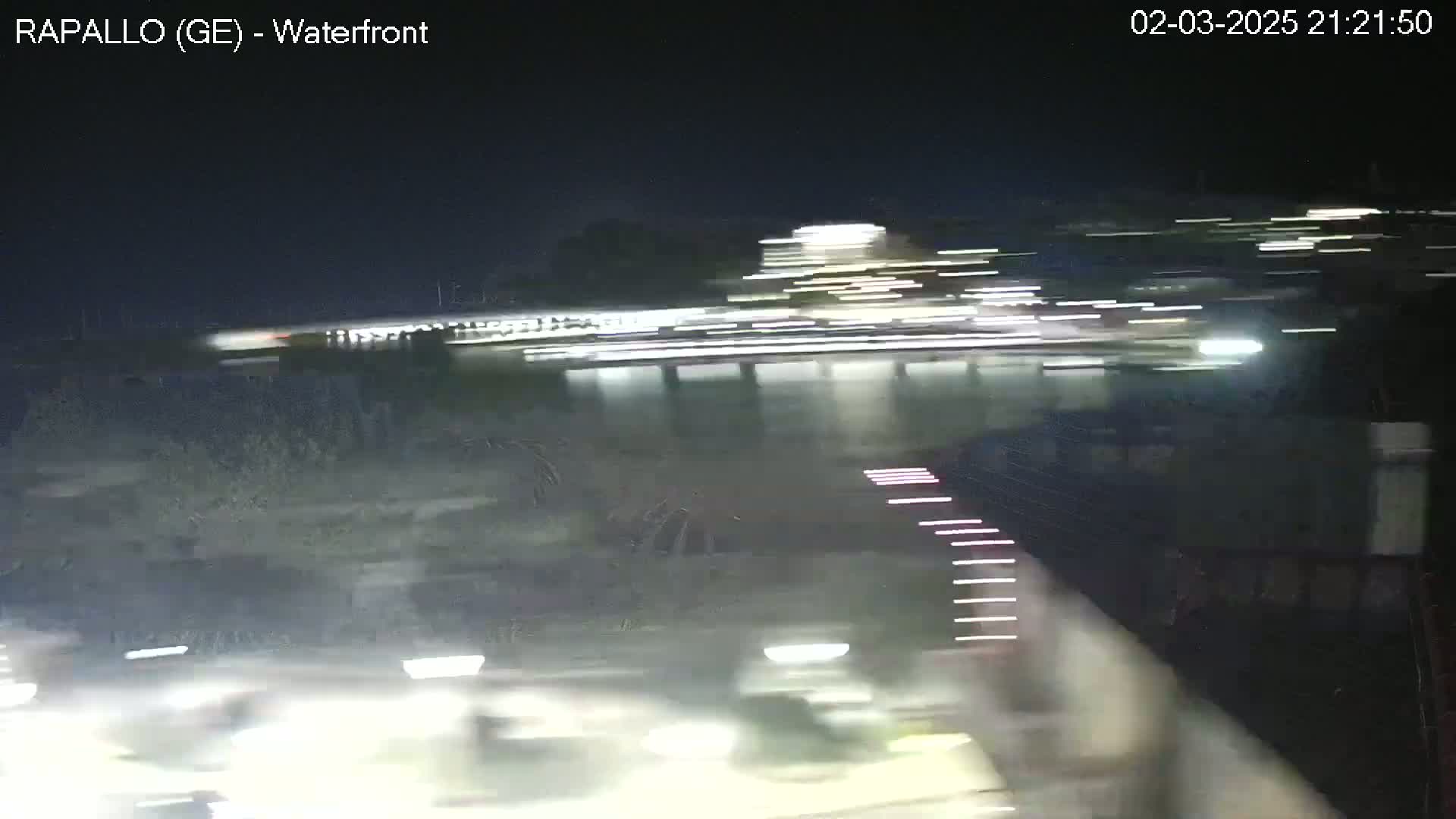 A nighttime waterfront scene shows blurred lights from moving vehicles and buildings across a dark body of water.