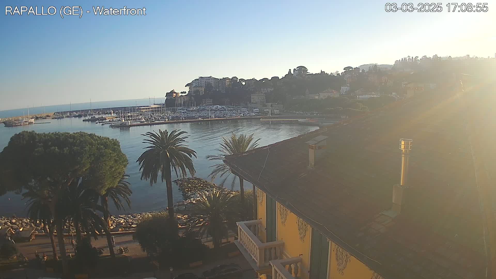 A waterfront view shows a harbor filled with boats, a hillside town, palm trees, and a building with a yellow facade under a clear, sunny sky.