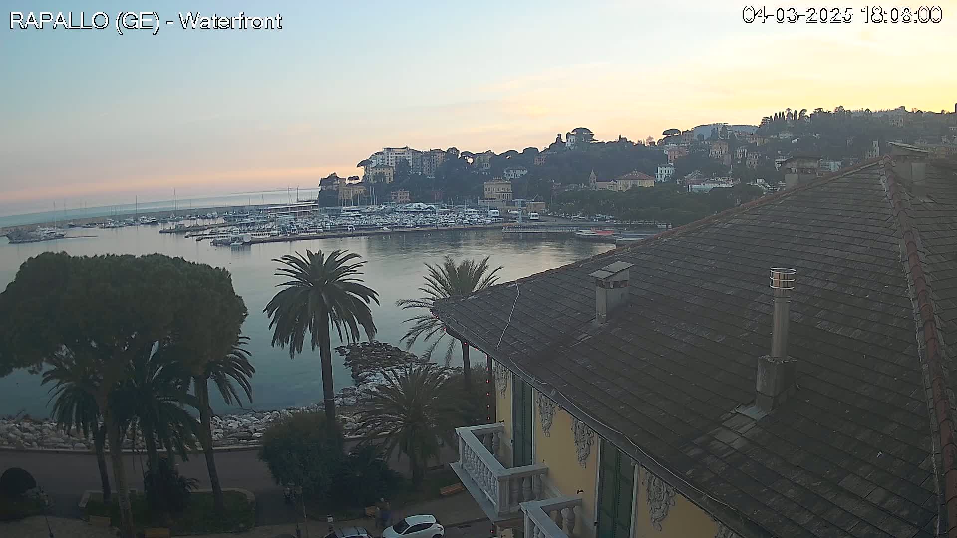 A waterfront view at sunset shows a harbor filled with boats, a hillside town, palm trees, and the roof of a building.