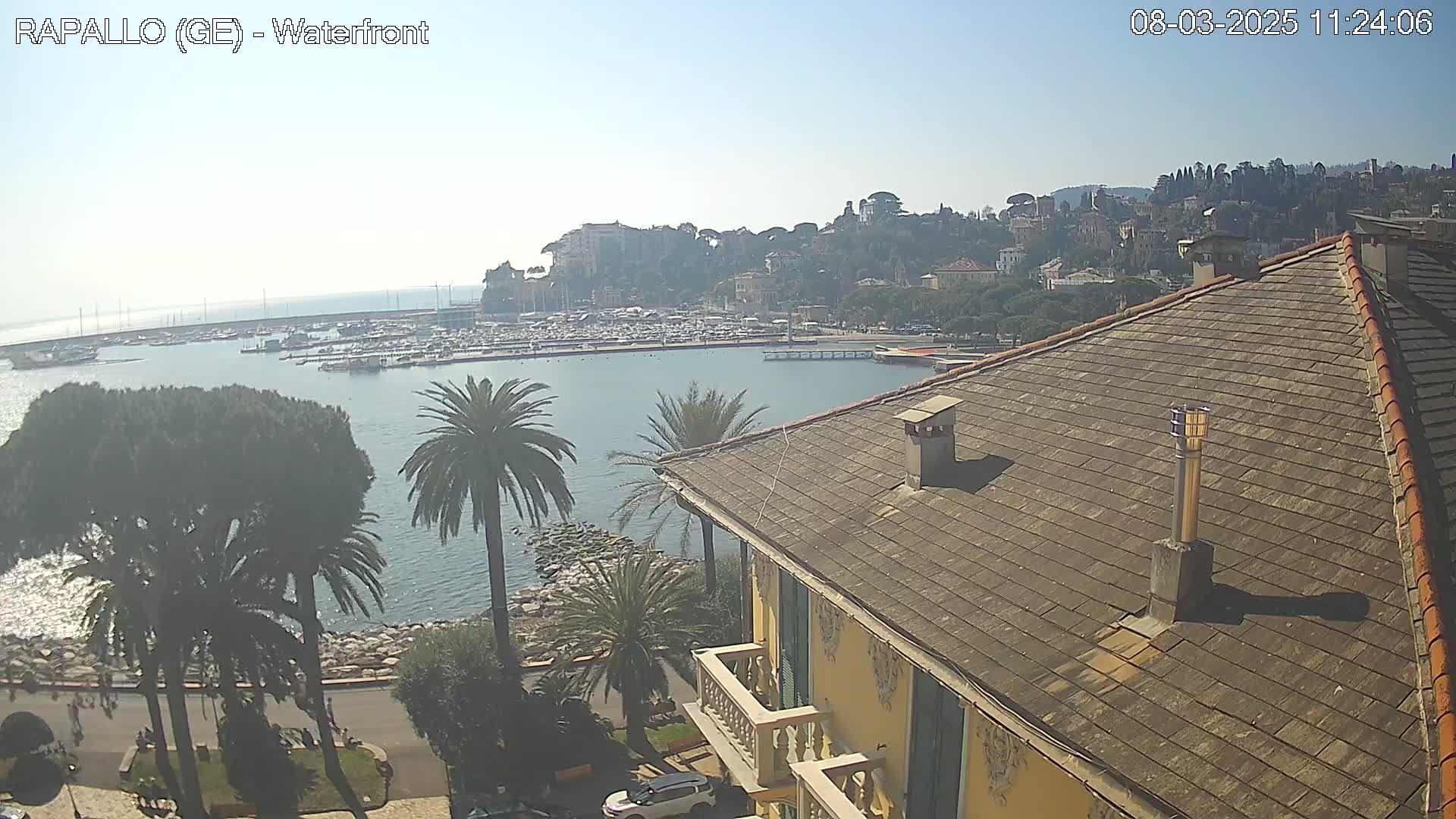 A waterfront view of a harbor filled with boats, a town on a hillside, and a portion of a building with a sloped roof, all under a clear, sunny sky.