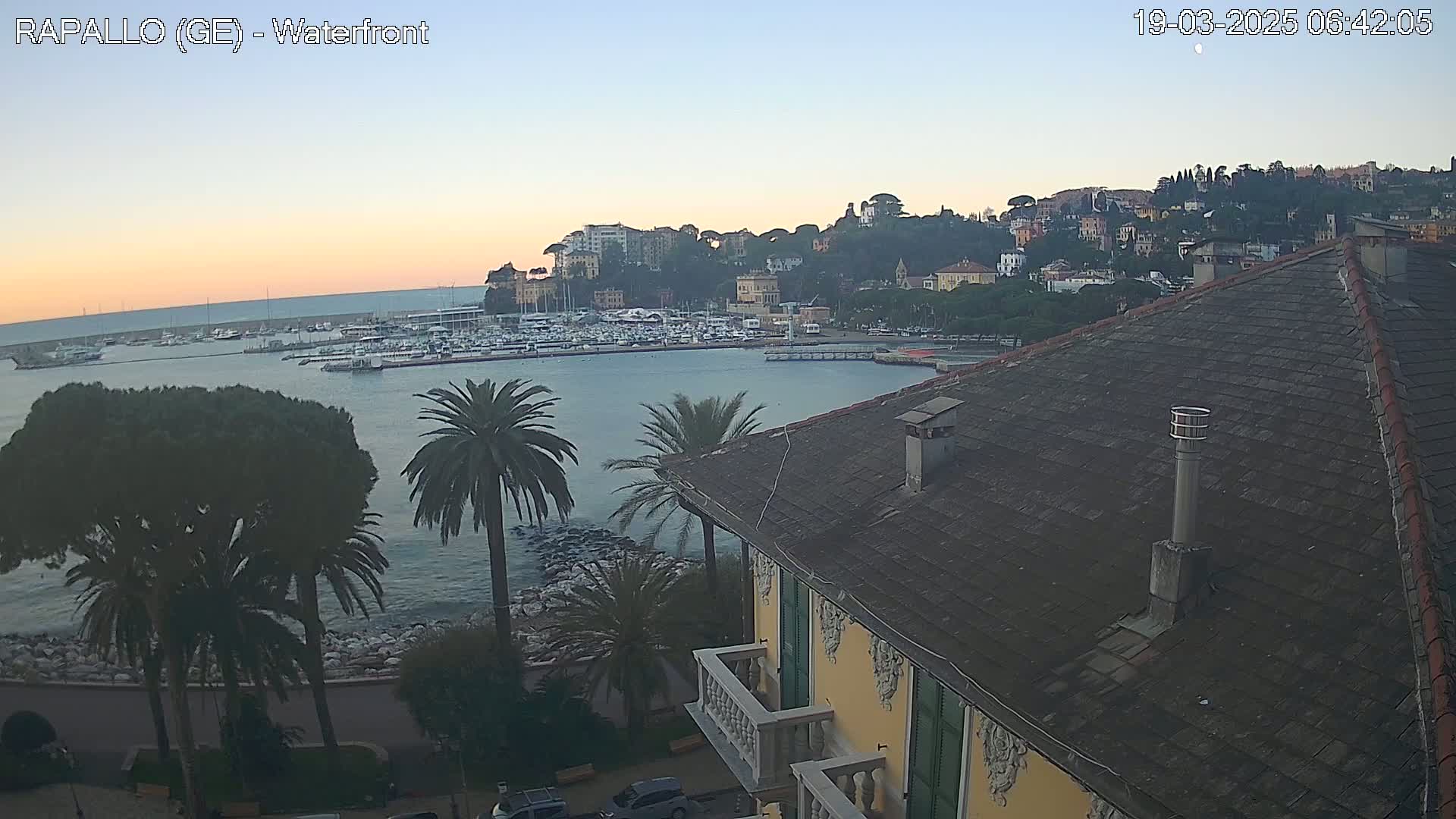 A waterfront view at dawn shows a calm harbor filled with boats, a hillside town with buildings nestled among trees, and a section of a building with a tiled roof in the foreground.