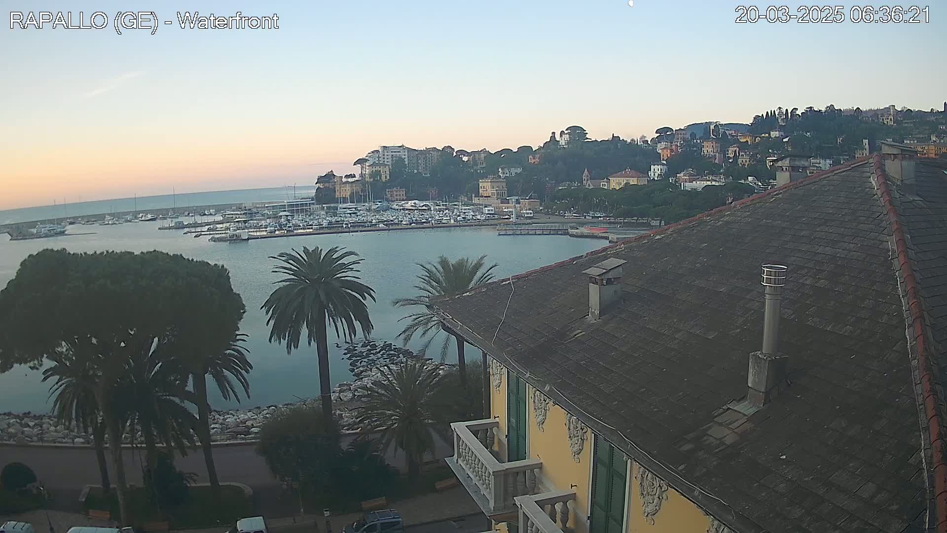 A waterfront view of a town nestled on a hillside overlooking a calm harbor filled with boats, under a twilight sky.