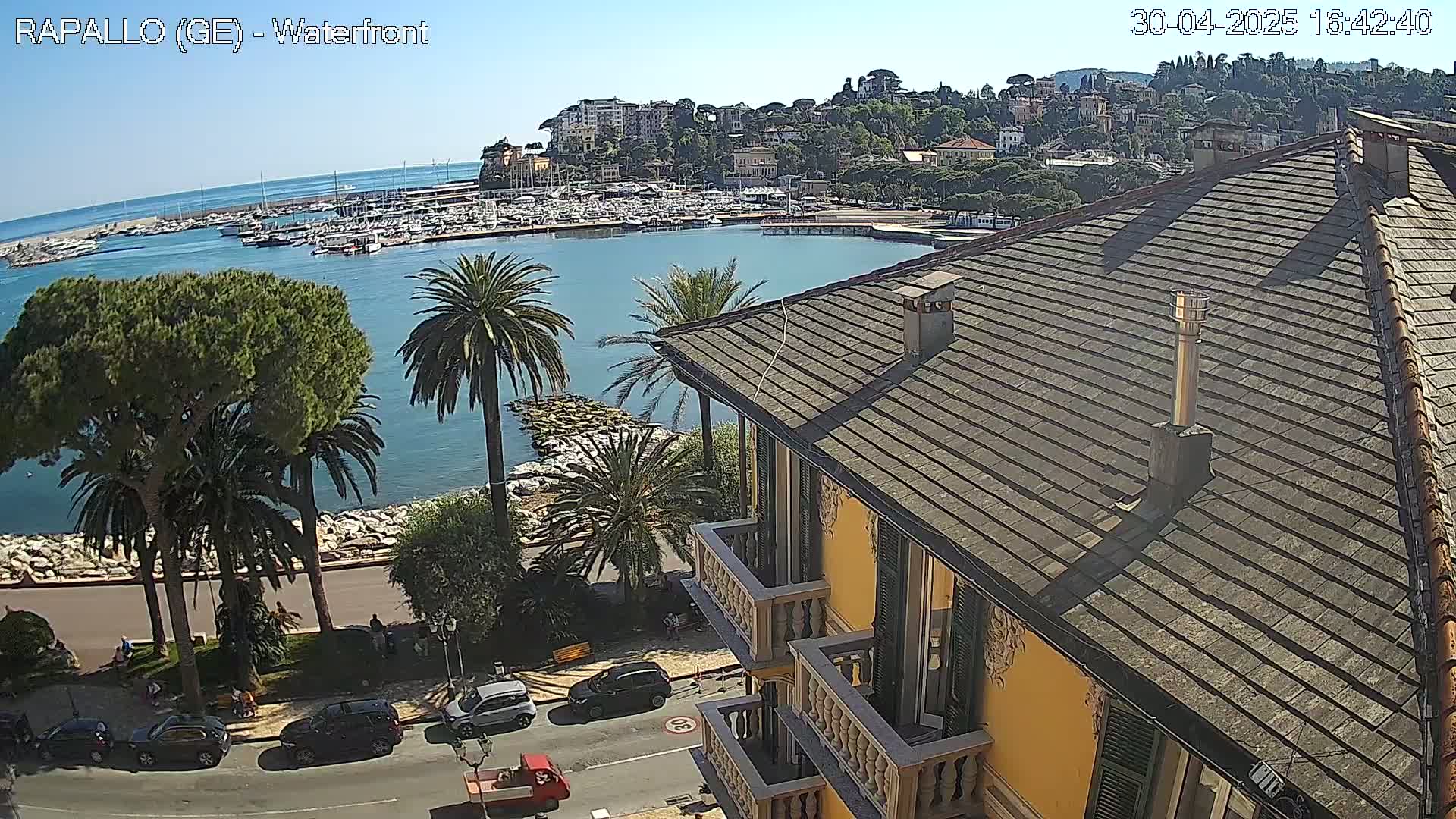 A sunny waterfront view shows a harbor filled with boats, a town nestled on a hillside, palm trees lining a seaside promenade, and a portion of a yellow building with a tiled roof in the foreground.
