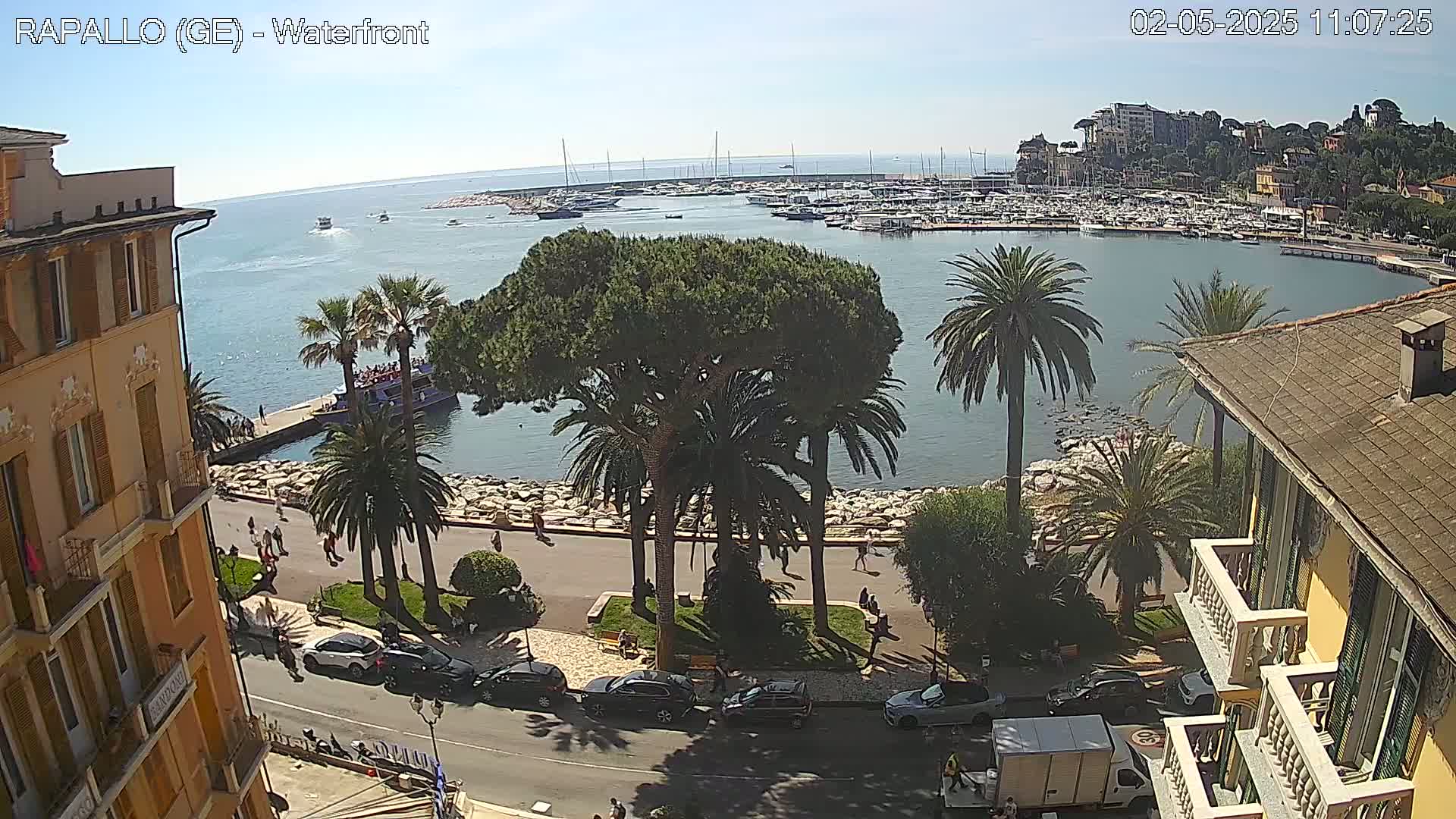 A waterfront scene on a sunny day shows a harbor filled with boats, palm trees lining a paved walkway, and buildings along the shore.
