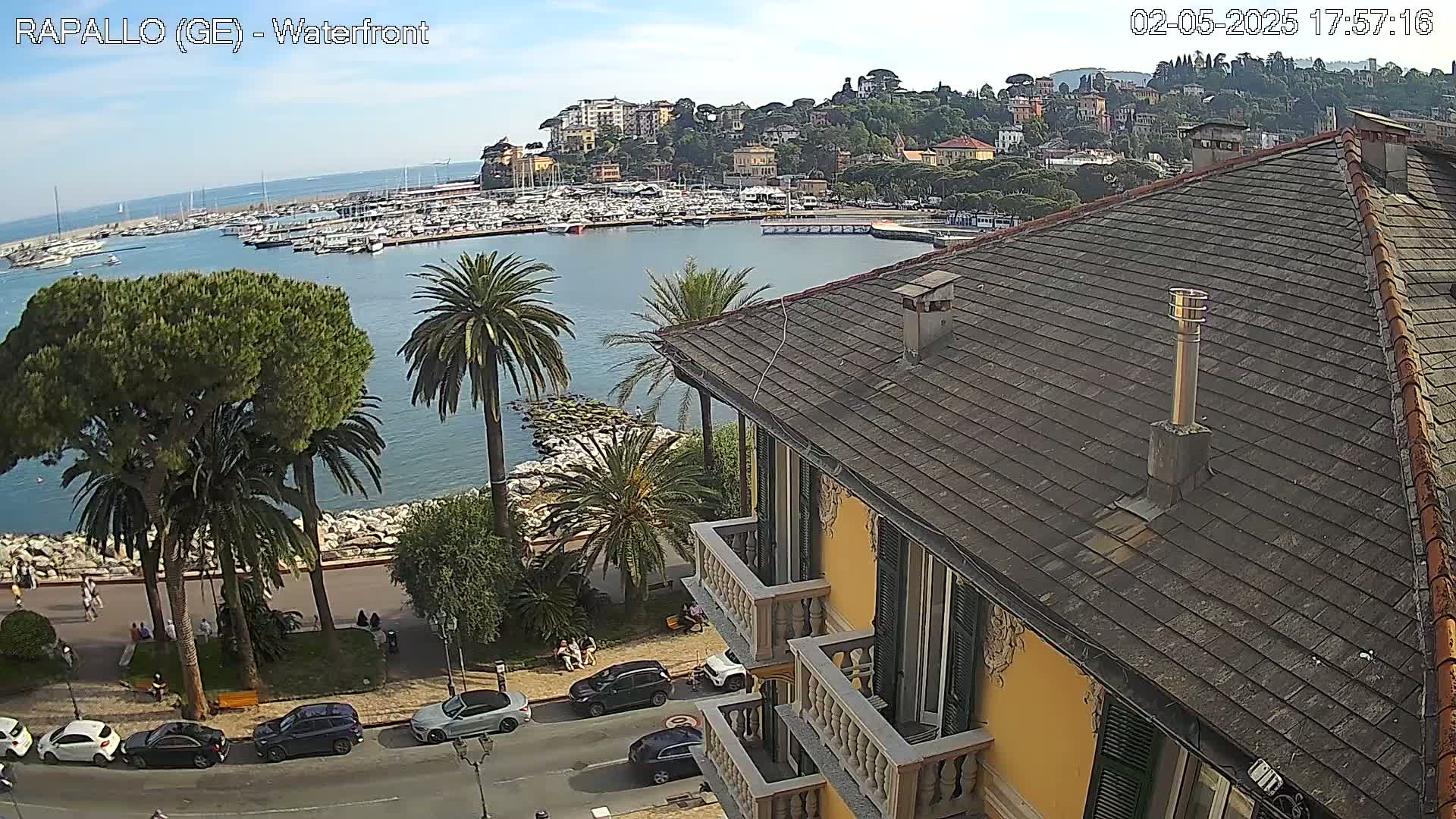 From a rooftop, a harbor filled with boats is visible beneath a partly sunny sky, with a town nestled on a hillside in the background.