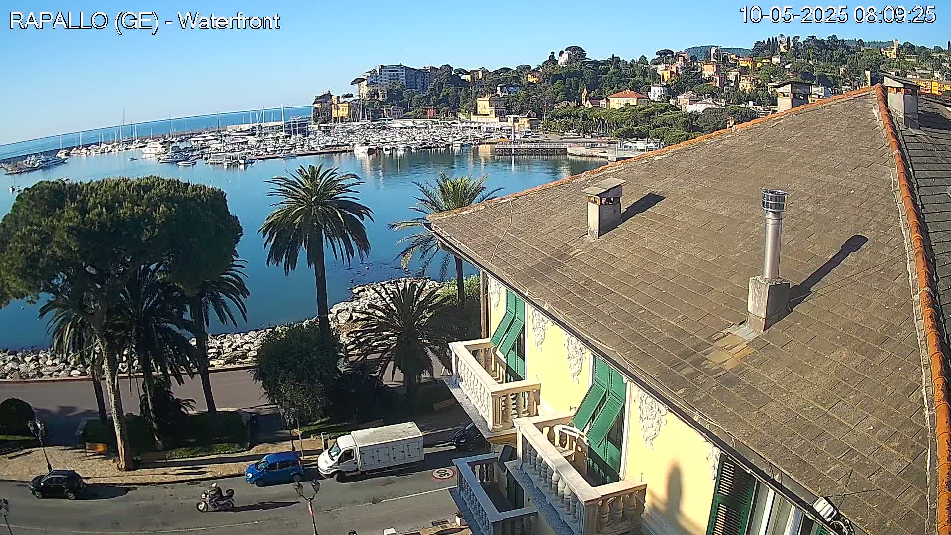 A waterfront view on a sunny day shows a harbor filled with boats, a town nestled on a hillside, and a portion of a yellow building with green shutters in the foreground.