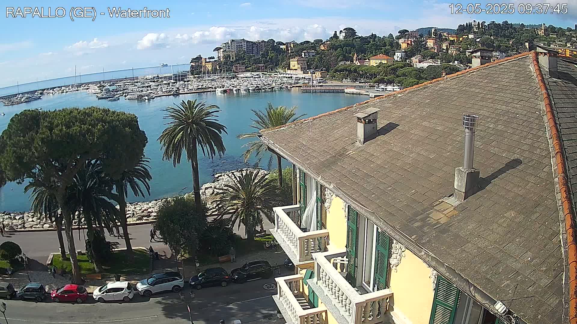 A waterfront view of a town with a harbor full of boats, under a mostly sunny sky, is seen from a rooftop of a multi-story building with balconies.