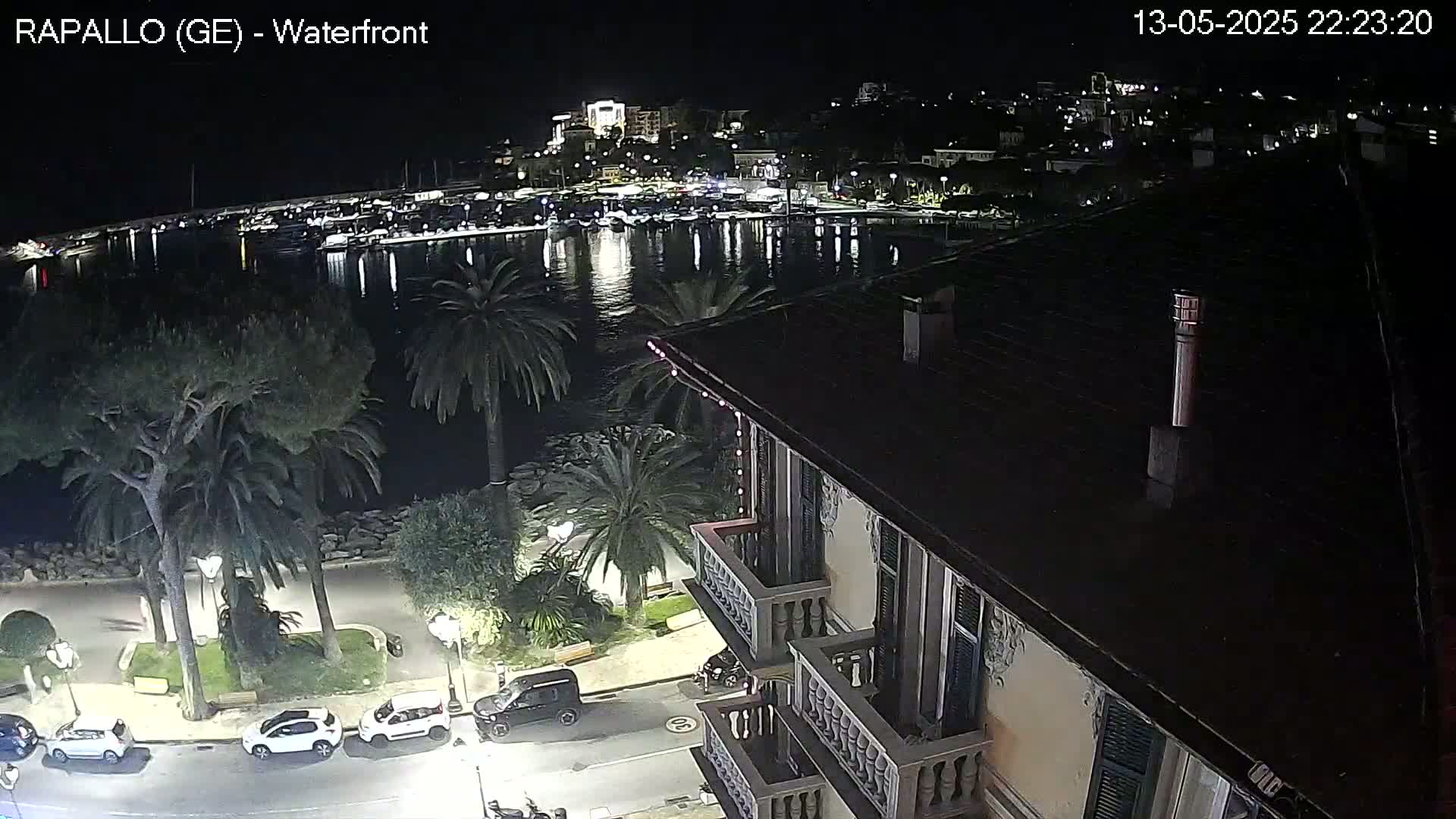 A nighttime view from a building overlooking a calm harbor filled with boats, a waterfront promenade with palm trees and parked cars, and a town lit up in the distance.