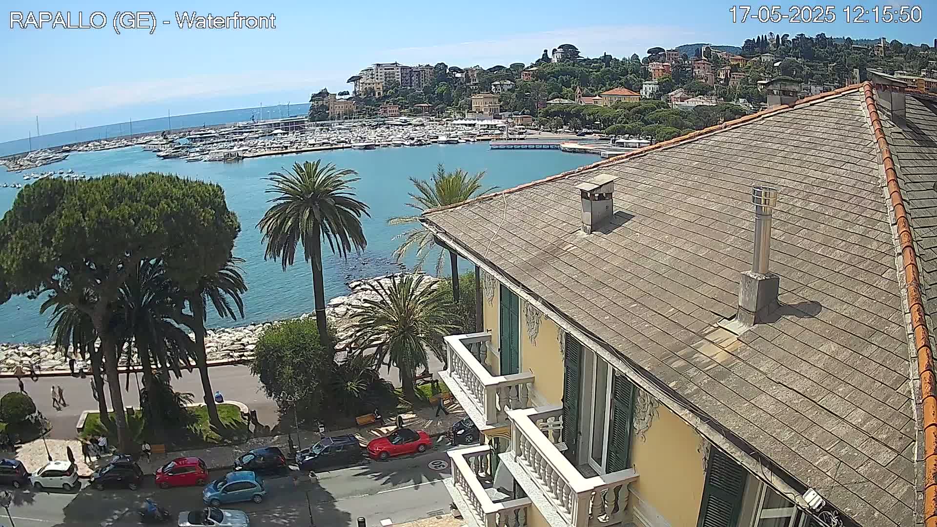 A waterfront view of a harbor filled with boats, a town nestled on a hillside, and a portion of a building with a sloped roof, all under a sunny sky.