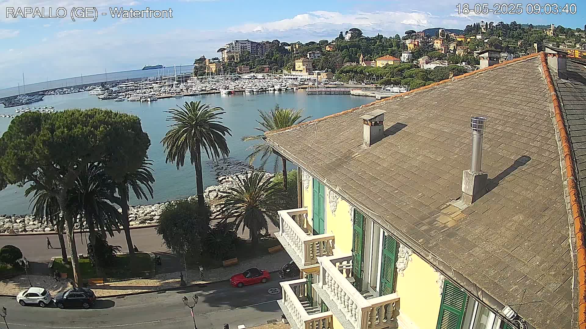 A partly sunny day overlooking a harbor filled with boats and a hillside town, viewed from a yellow building's rooftop.