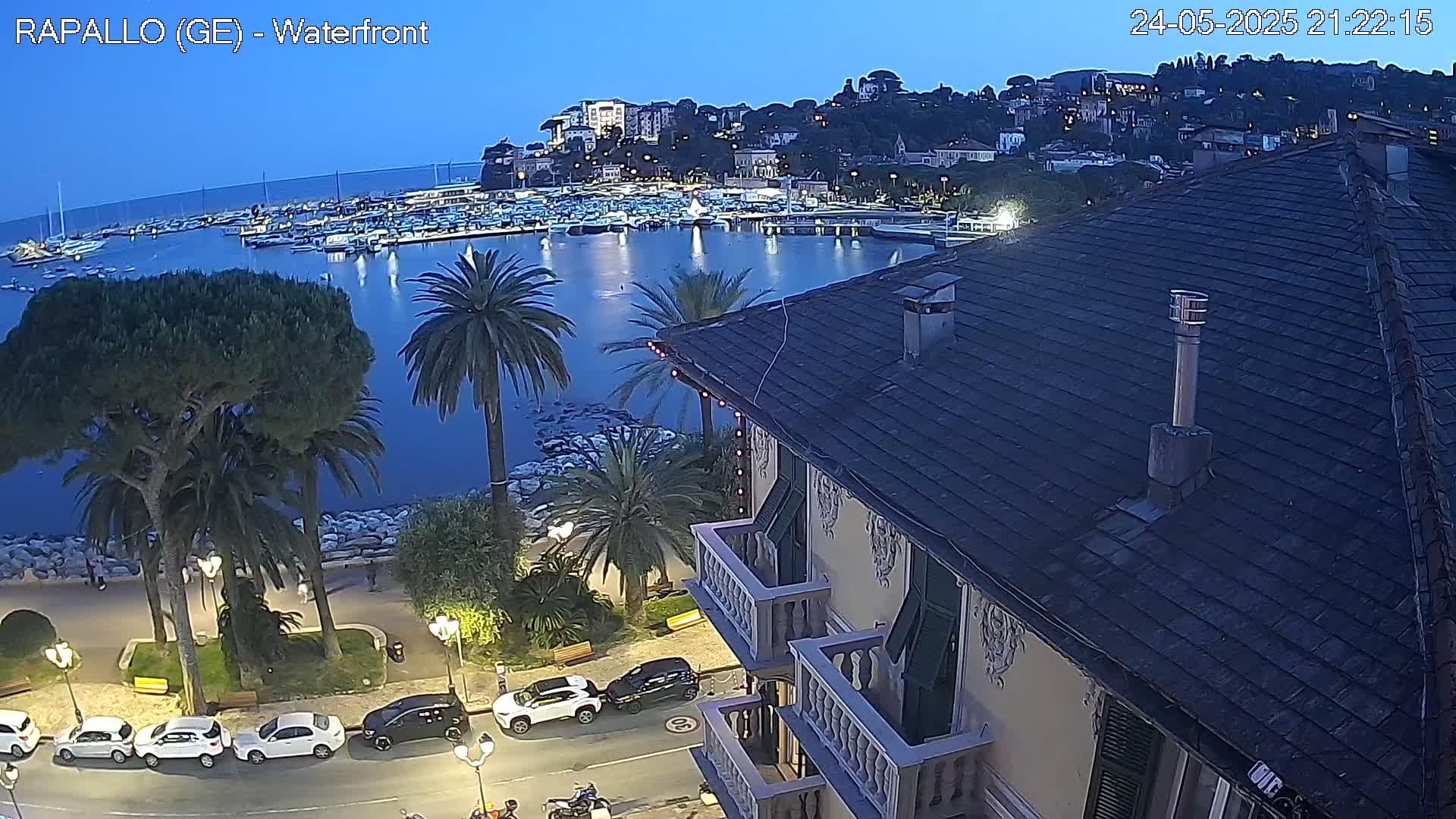 An evening view of a harbor filled with boats, a town nestled on a hill, and a seaside promenade with palm trees, seen from an elevated vantage point overlooking a building with balconies.