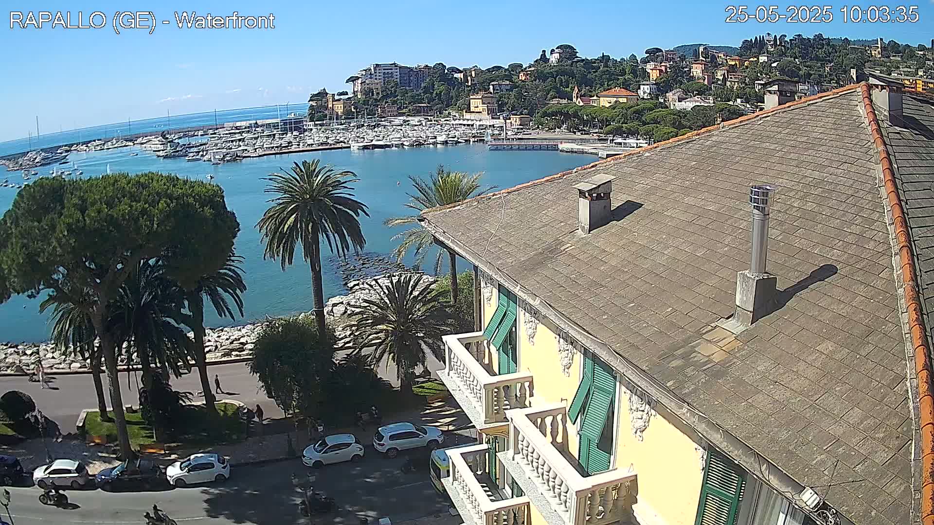 A waterfront view on a sunny day shows a harbor filled with boats, a town nestled on a hillside, and a yellow building with balconies in the foreground.