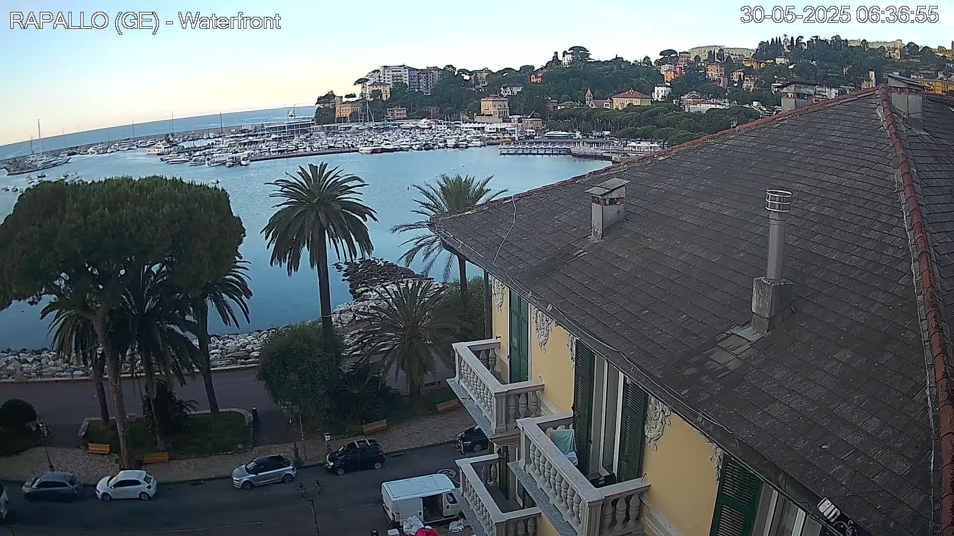 A view from a building overlooking a calm harbor filled with boats, a seaside town nestled on a hillside, and a street with parked cars, all under a clear sky.