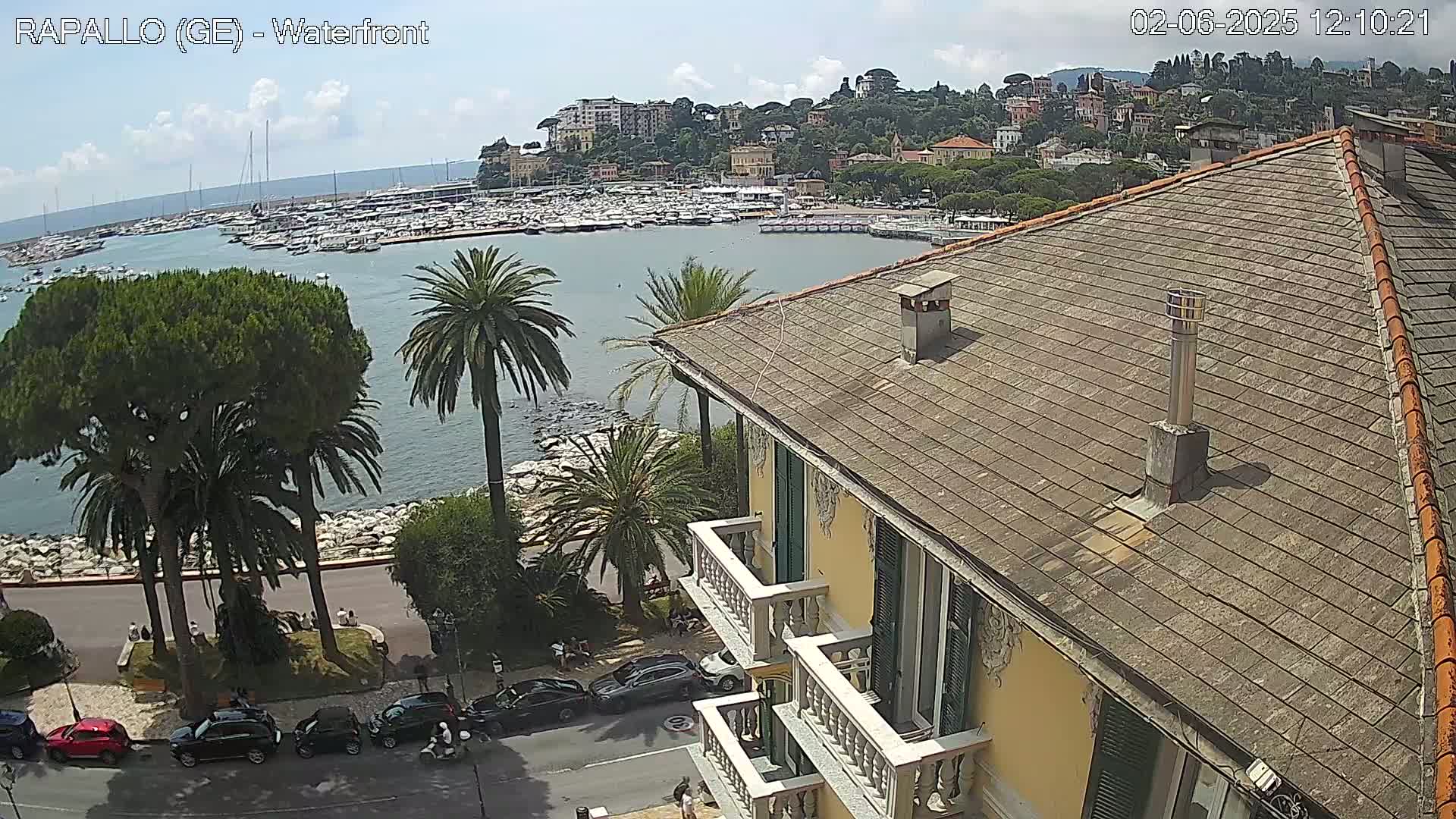 A partly sunny waterfront view shows a harbor filled with boats, a town nestled on a hillside, palm trees, and cars parked along a street below a yellow building with a gray roof and balconies.