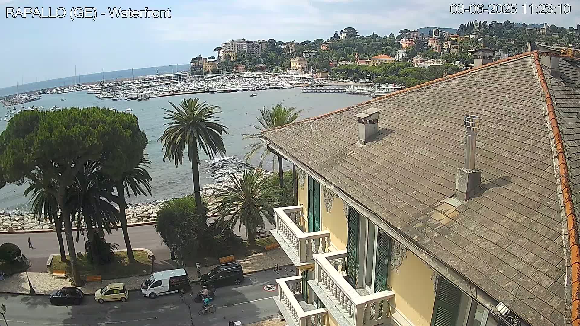 A partly sunny day overlooks a harbor filled with boats, a seaside town nestled against a hillside, and a building's rooftop with balconies.
