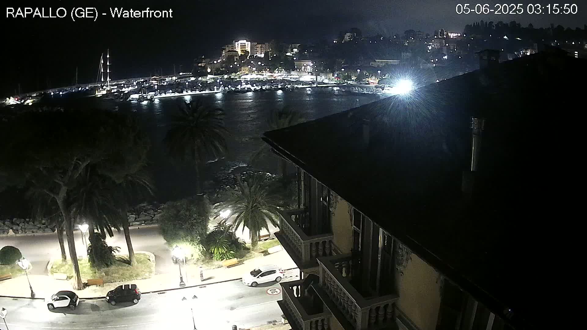 A nighttime view from a building overlooking a calm harbor filled with boats, a waterfront lined with palm trees, and a city illuminated in the distance.