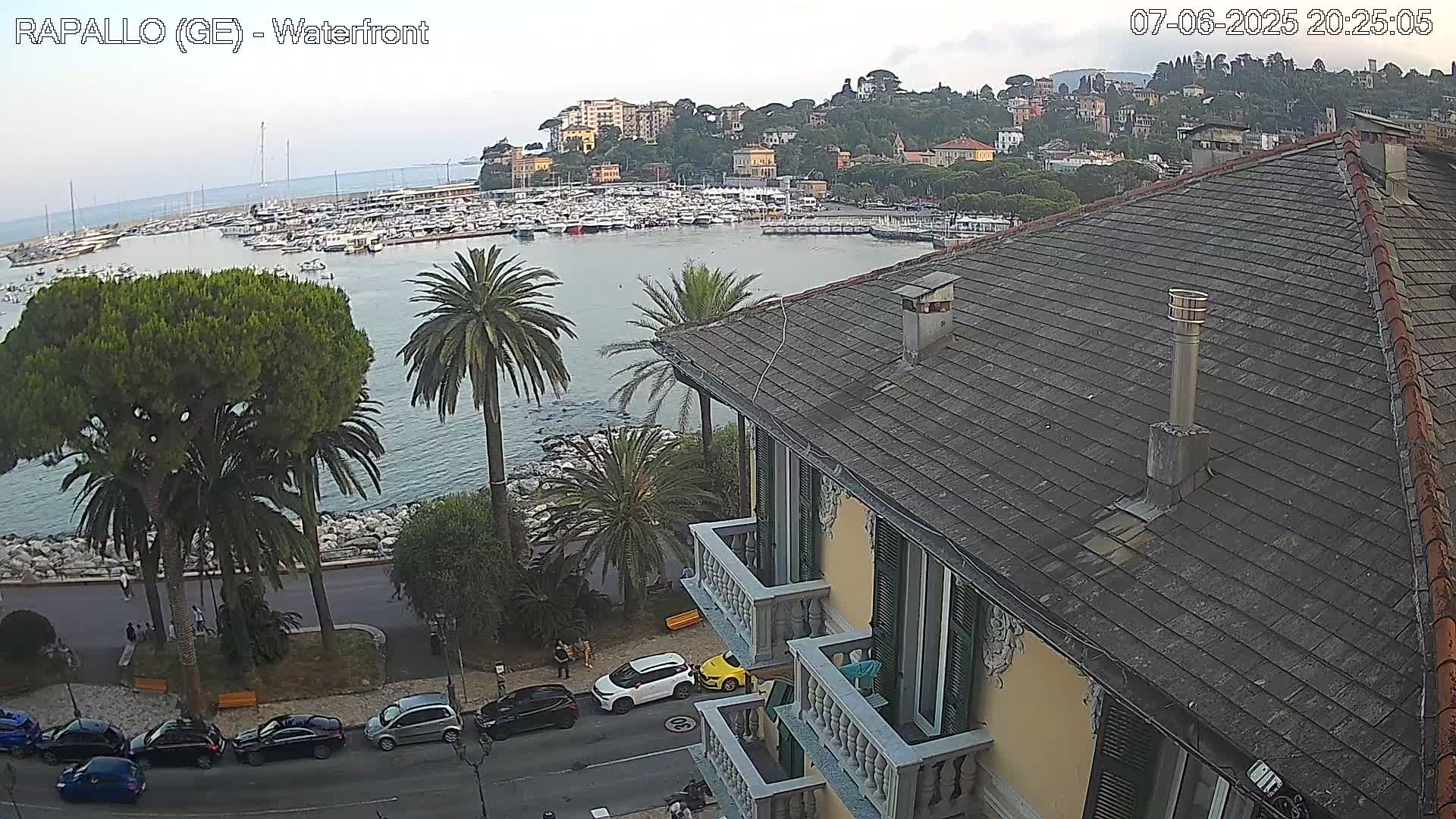 An overlook of a harbor filled with boats, a town on a hillside, and a seaside road with cars parked along it, all under a mostly clear sky.