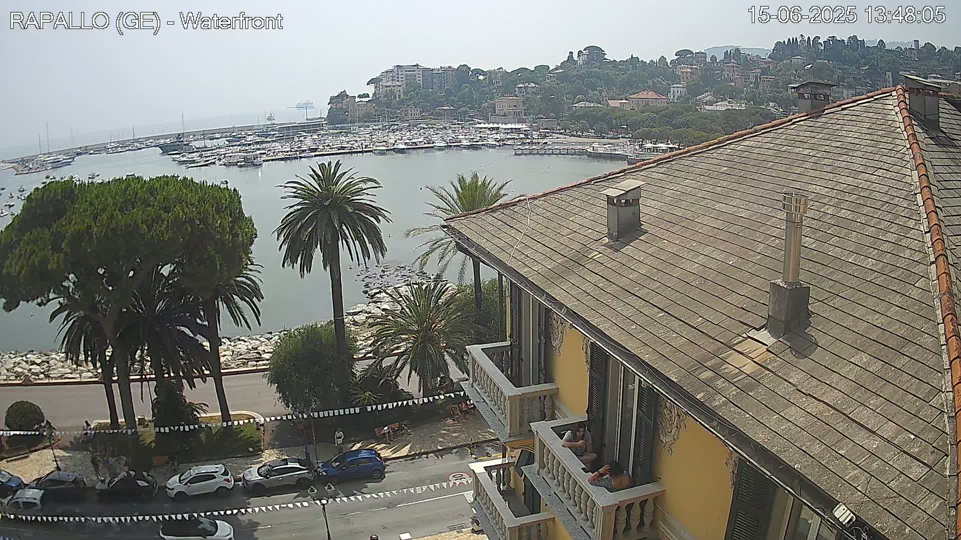 A hazy view of a harbor filled with boats, a town nestled on a hillside, and a building with people on its balcony, all under a sunny sky.
