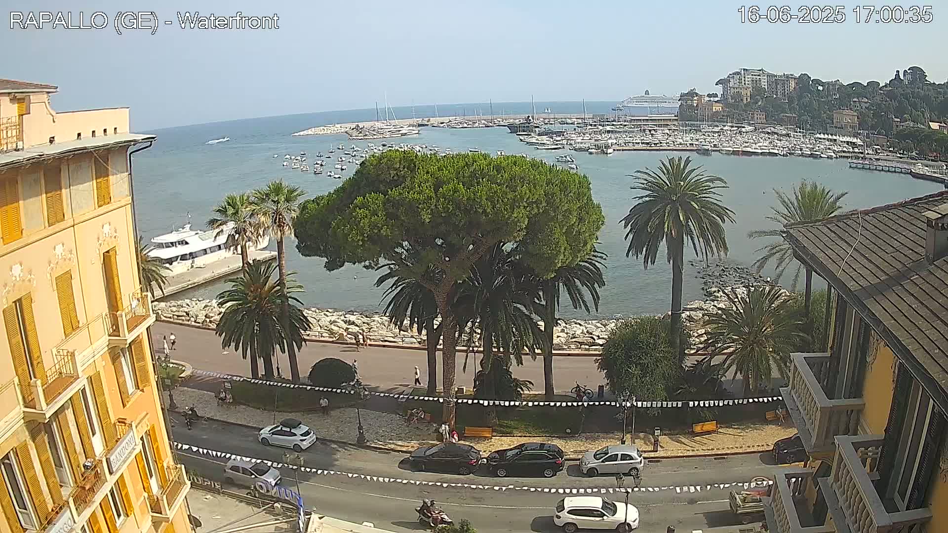 A sunny waterfront scene shows a harbor filled with boats, palm trees lining a paved walkway, and buildings along a seaside road.