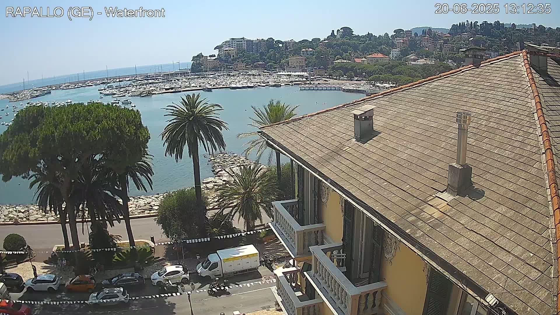 A waterfront view of a harbor filled with boats, a town nestled on a hillside, and palm trees lining a roadway, all under a clear, sunny sky, is seen from a rooftop.
