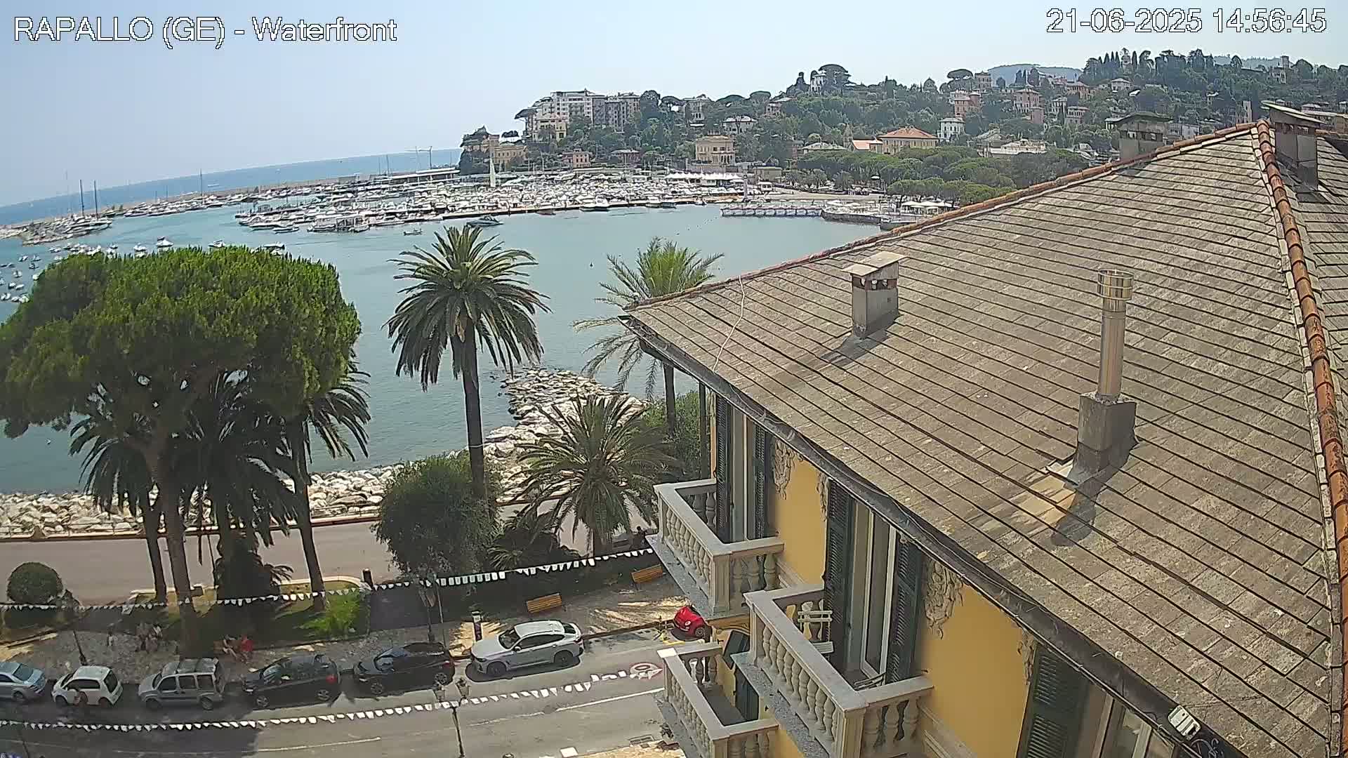A waterfront view of a harbor filled with boats, a town nestled on a hillside, and a building with a sloped roof, all under a clear, sunny sky.
