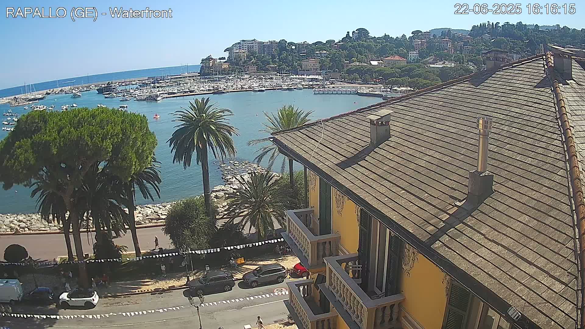 A waterfront scene on a sunny day shows a harbor filled with boats, a town nestled on a hillside, palm trees lining a roadway, and a yellow building with a tiled roof in the foreground.