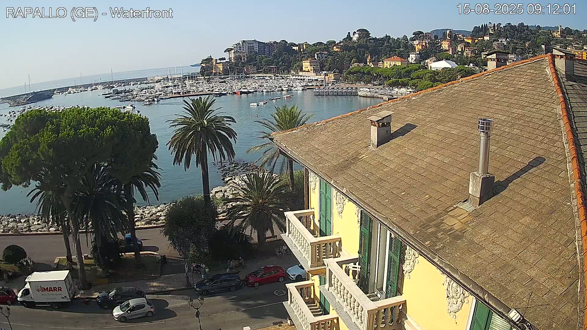 A high-angle view on a sunny day shows a harbor filled with boats, a town built on a hillside, and a yellow building with balconies overlooking a roadway and the water.