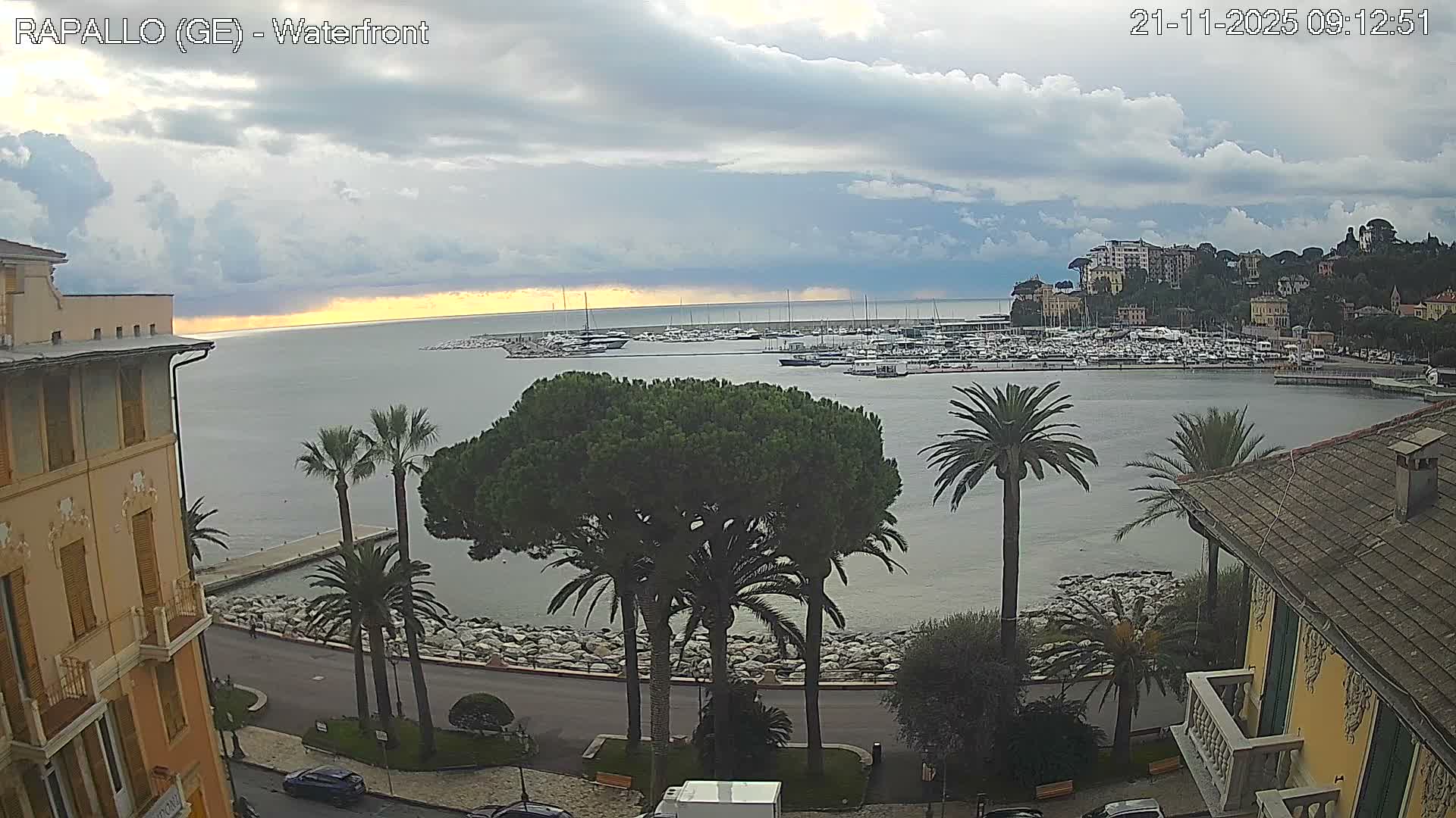 An expansive view captures a bustling coastal bay filled with numerous yachts and boats, bordered by a waterfront road, palm trees, and buildings, all under a heavily overcast sky with a band of distant golden light on the horizon.