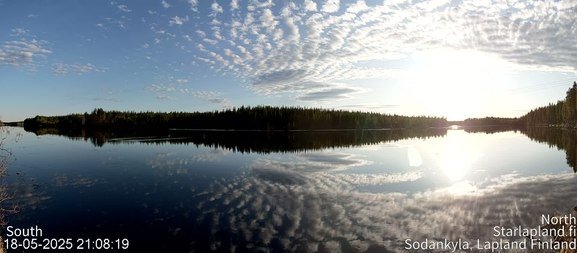 A calm lake reflects a mostly sunny sky filled with scattered clouds, bordered by a dark line of trees.