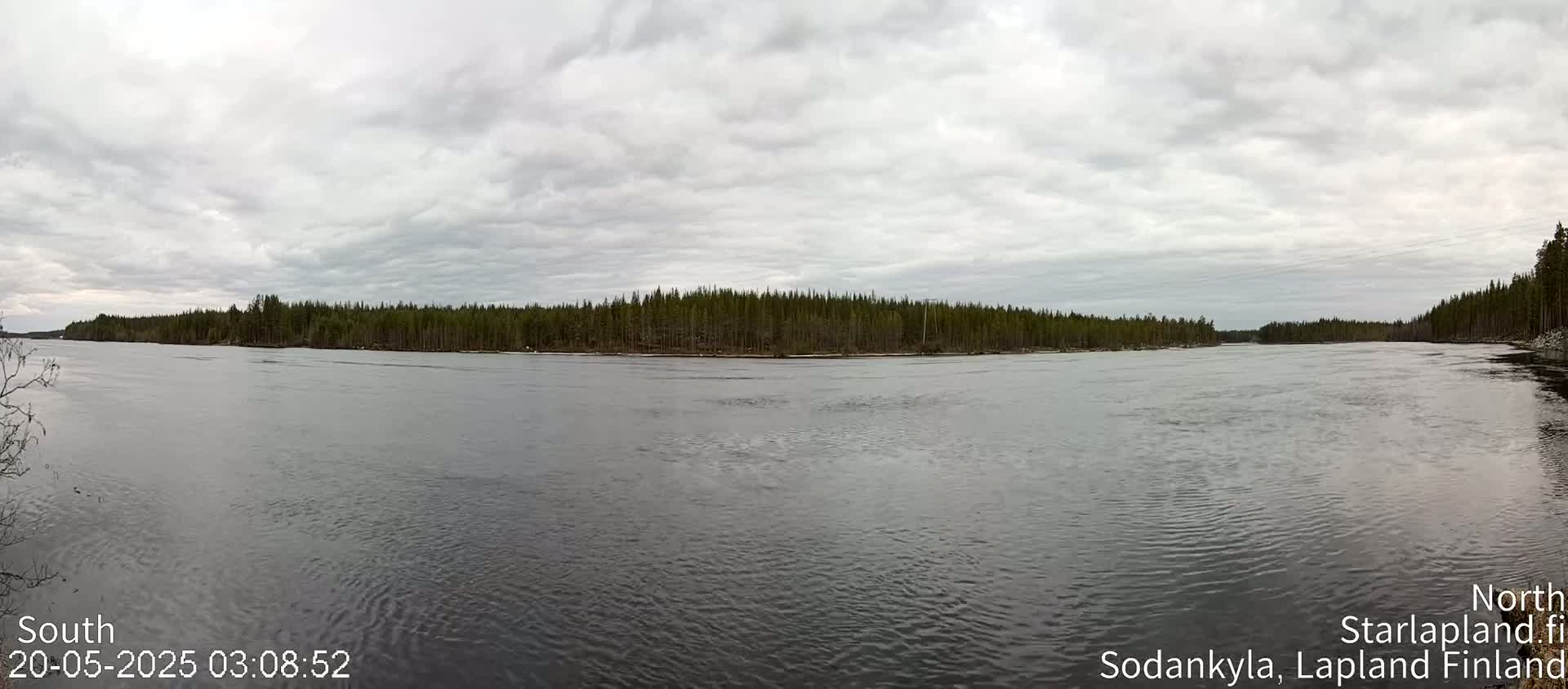 A wide, calm river flows under a cloudy sky, bordered by a dense line of evergreen trees.