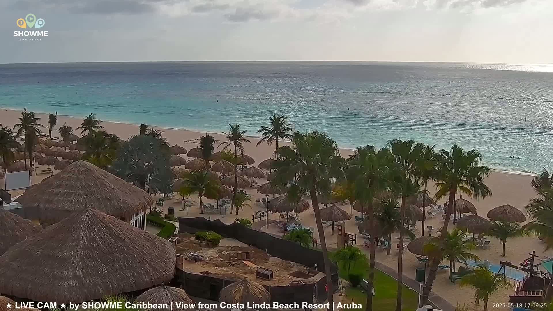 A beach with calm, light-blue water, numerous thatched umbrellas, and palm trees under an overcast sky.