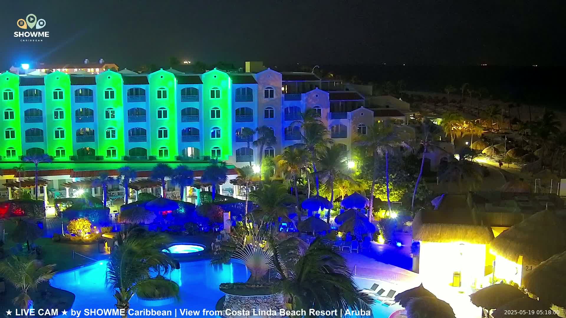 A brightly lit resort at night, featuring a multi-story building with green lighting, palm trees, pools, and thatched-roof structures.