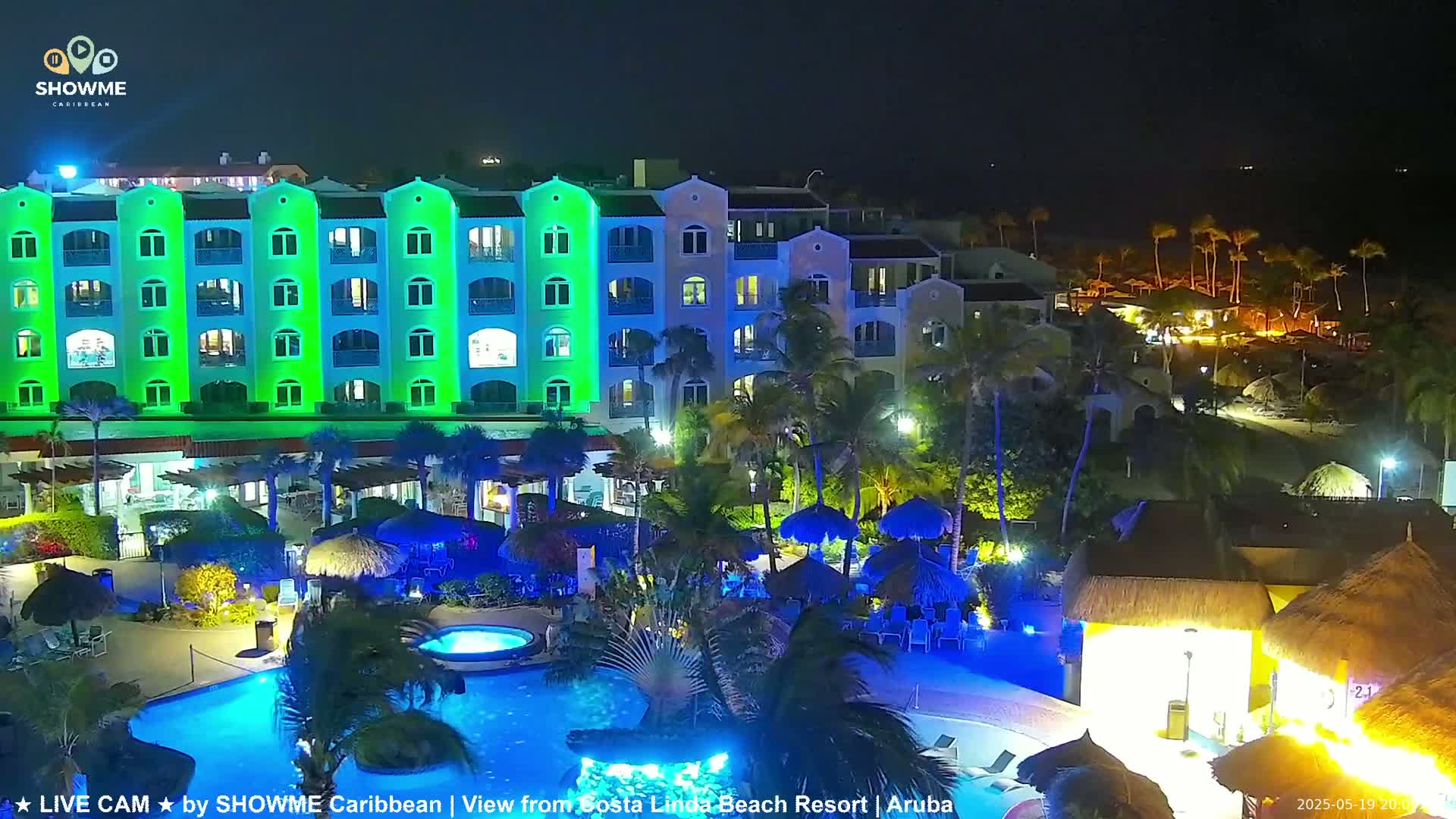 A brightly lit resort at night, featuring a swimming pool and a multi-story building with green and blue exterior lights, is surrounded by palm trees.