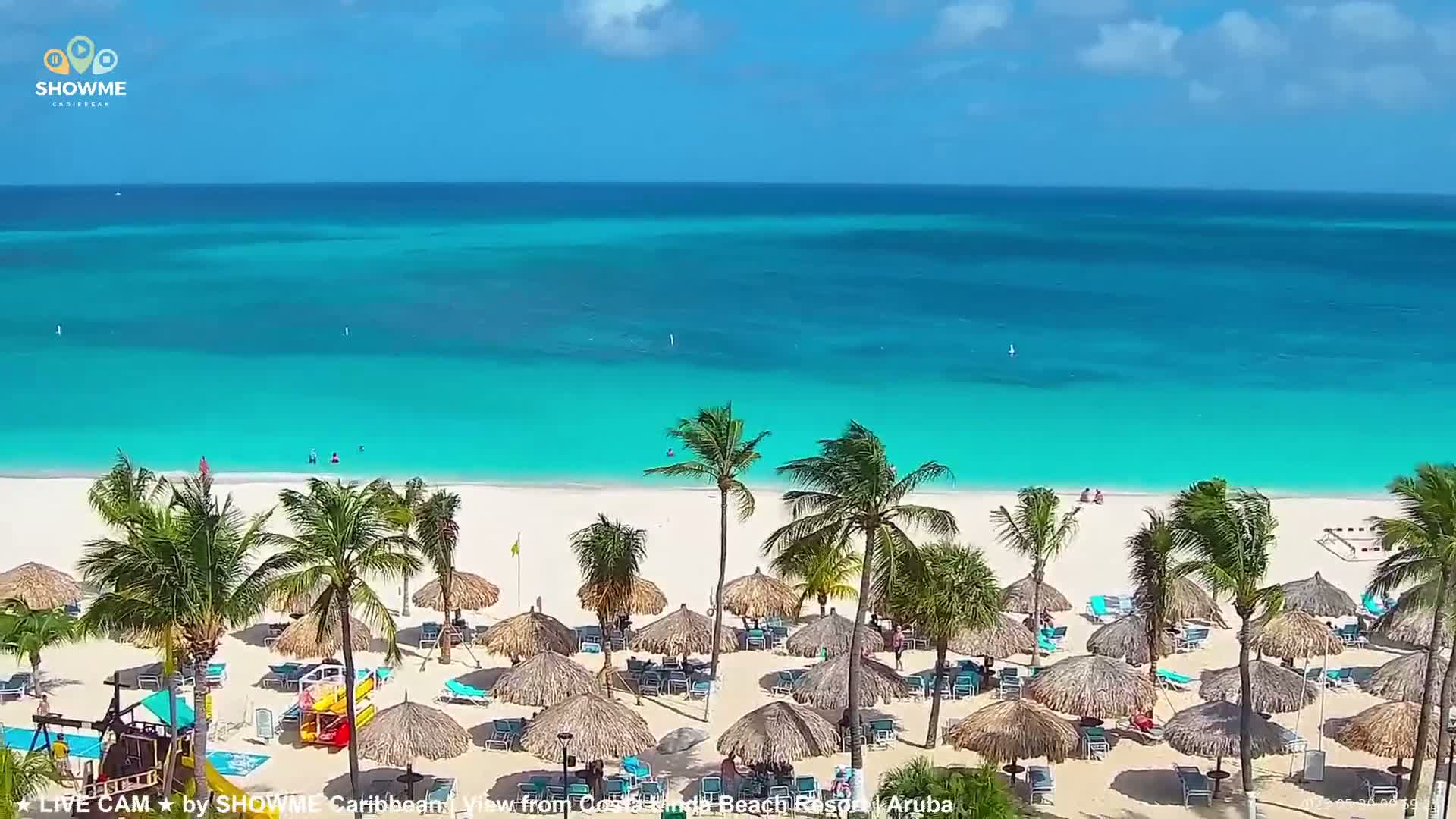 A white sand beach with turquoise water, numerous thatched umbrellas, and palm trees is shown on a sunny day.