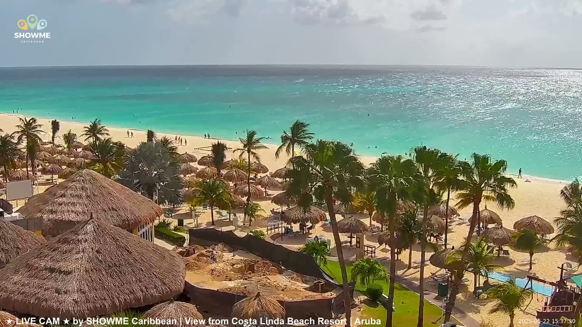 A beach with turquoise water, palm trees, and thatch-roofed umbrellas under a partly sunny sky.