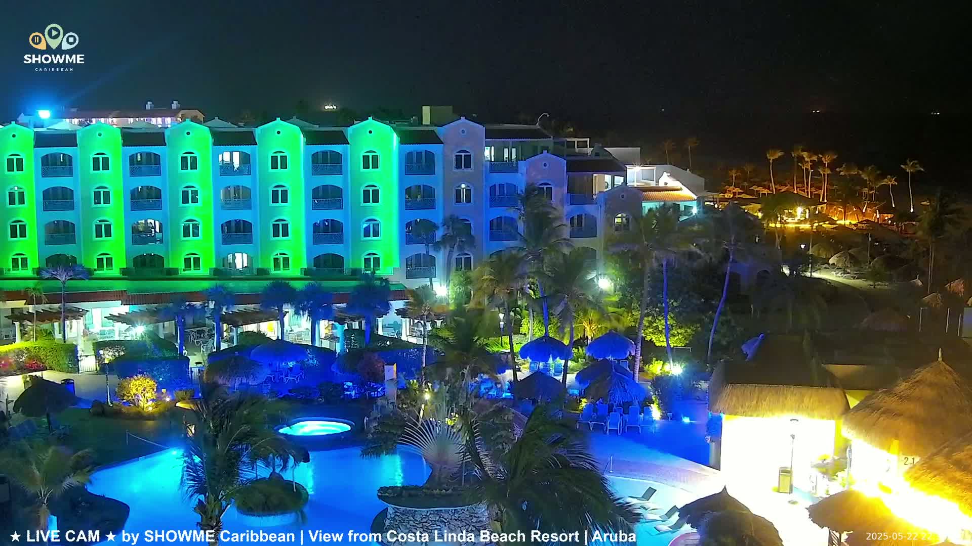 A brightly lit resort at night, featuring a multi-story building with green and blue lighting, a pool, and palm trees.