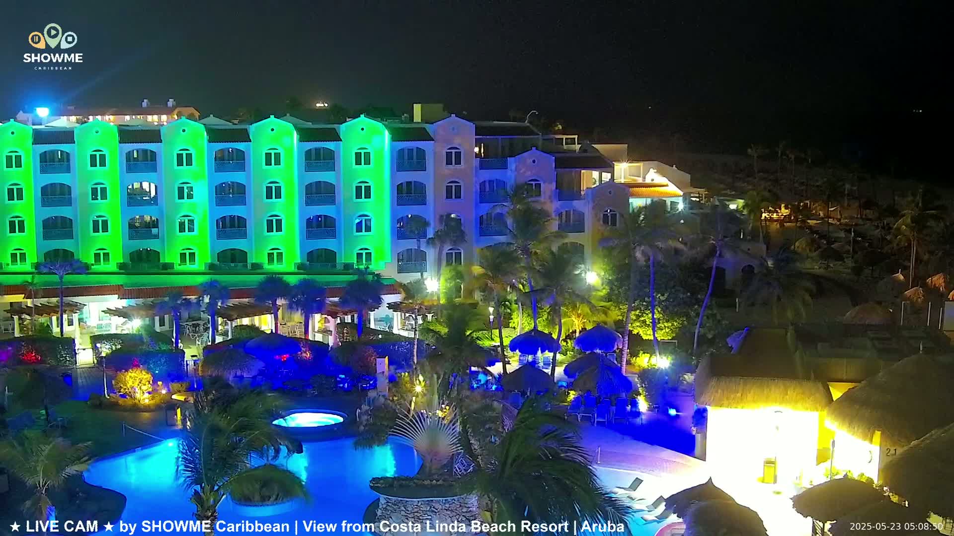 A brightly lit resort at night, featuring a multi-story building with green and blue lighting illuminating its exterior, along with a swimming pool and surrounding palm trees.