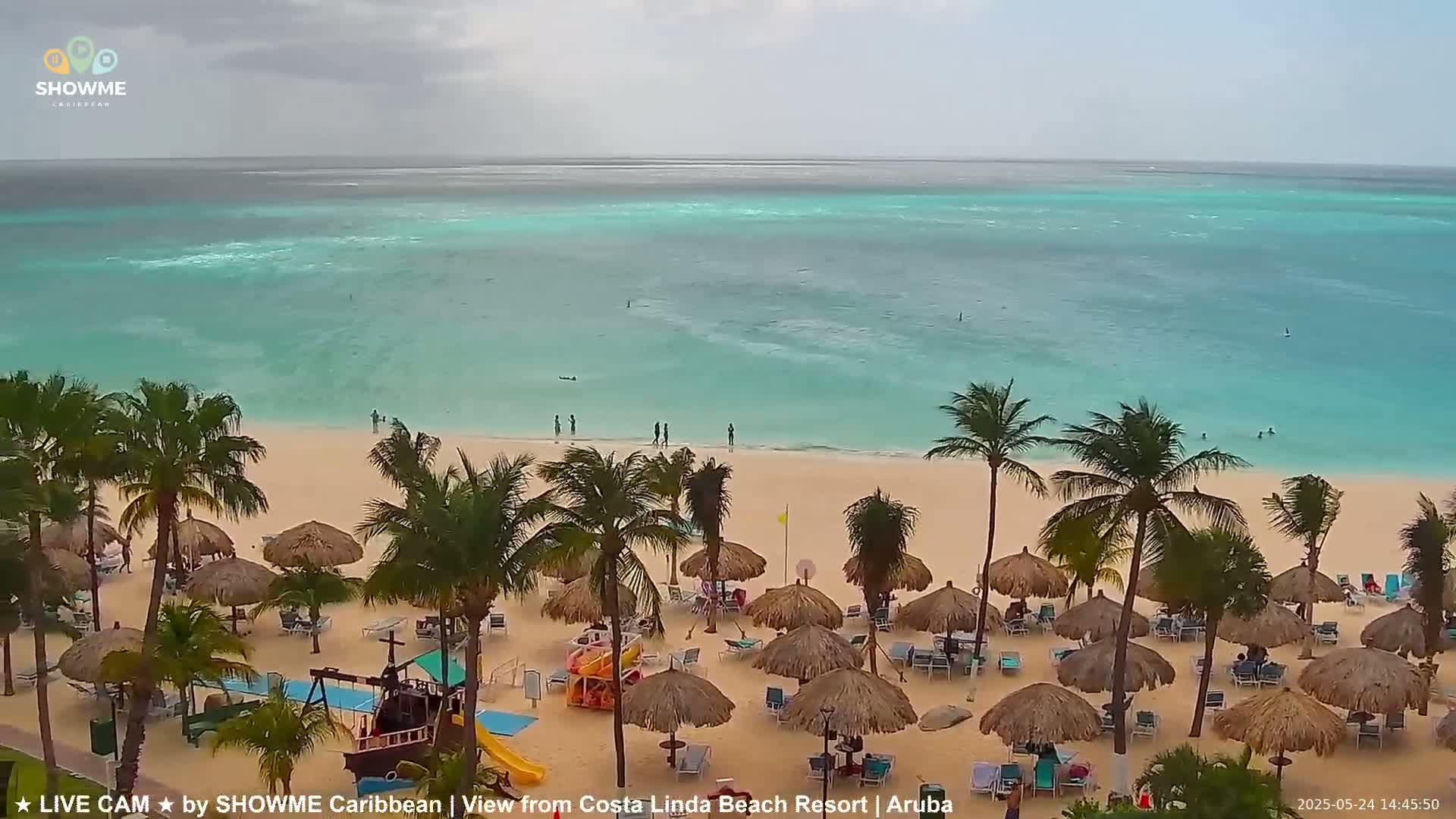 A beach with palm trees and thatched umbrellas under an overcast sky, showing a calm turquoise ocean in the background.