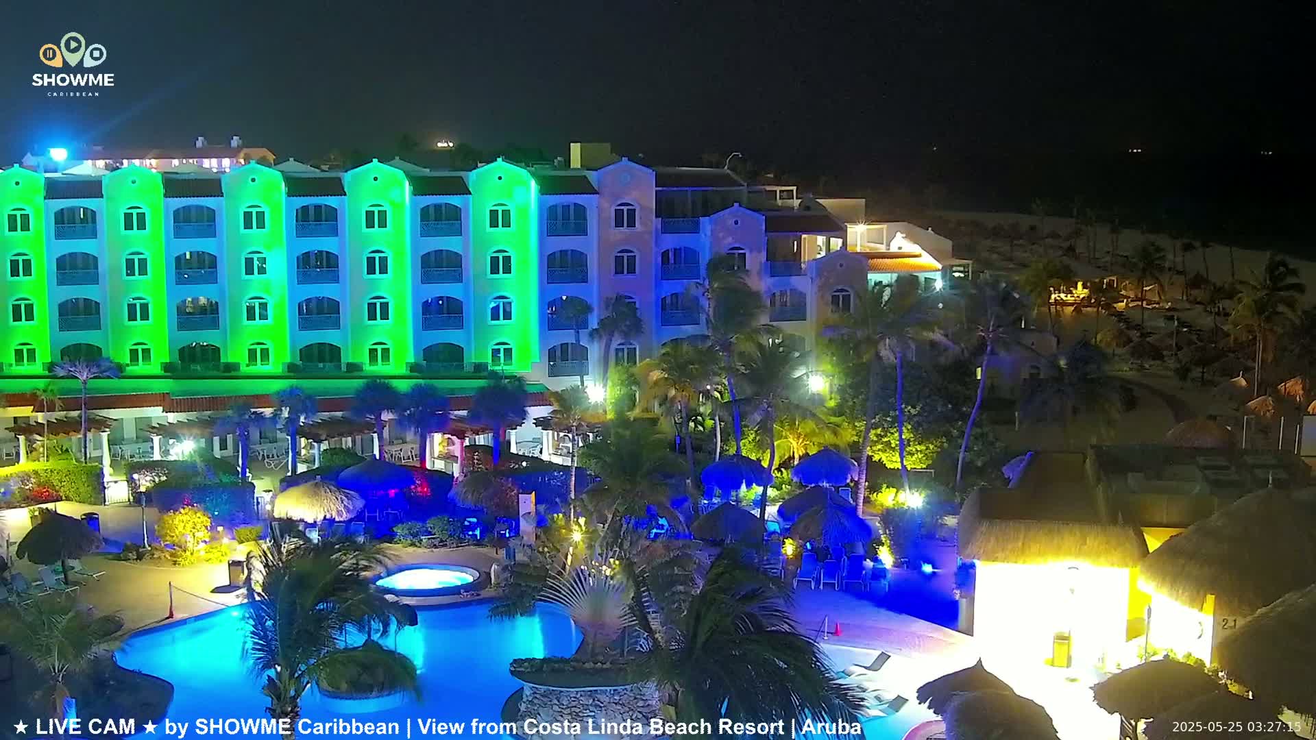 A brightly lit resort at night, featuring a large pool, palm trees, and a multi-story building illuminated in green and blue.
